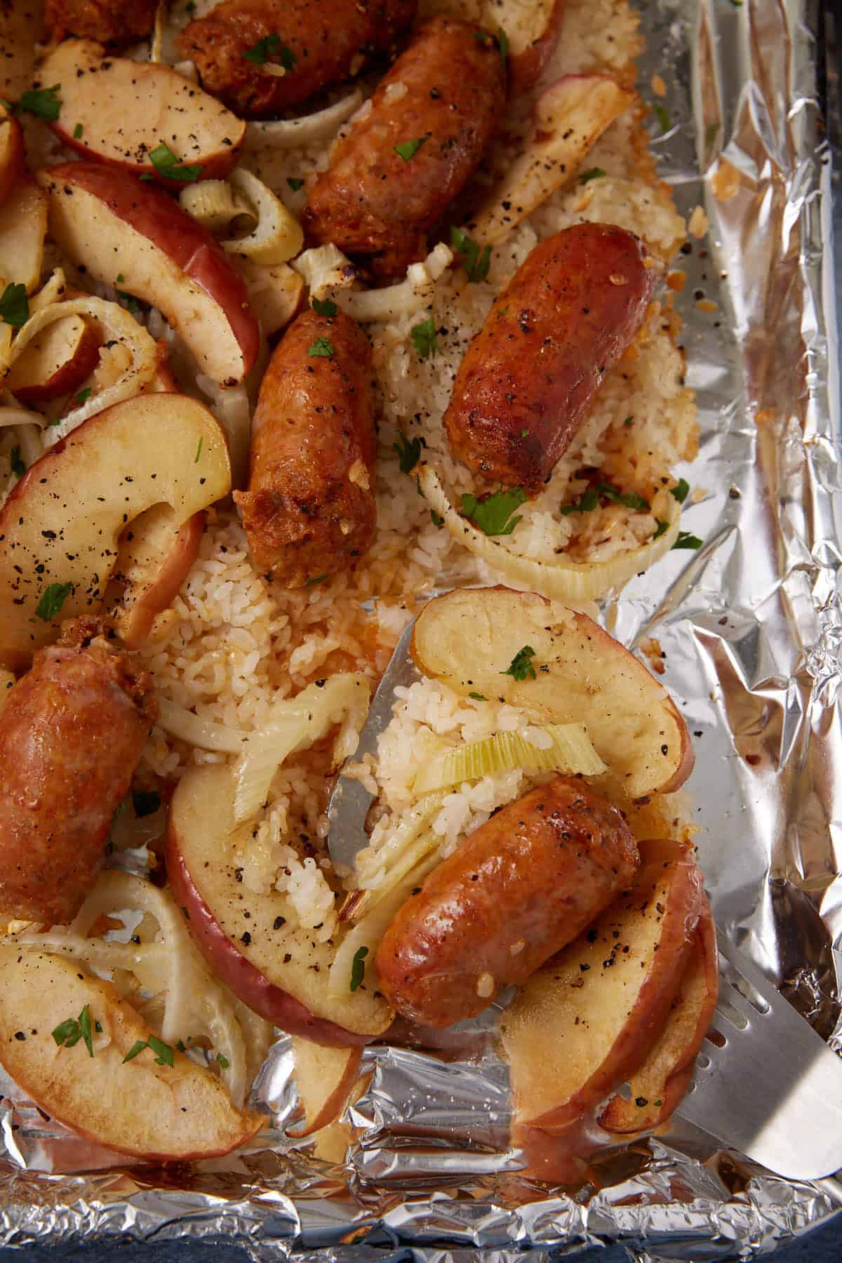 This sausage sheet pan dinner features sliced sausages, apple wedges, onions, and rice baked on a foil-lined tray, then garnished with chopped herbs and black pepper.
