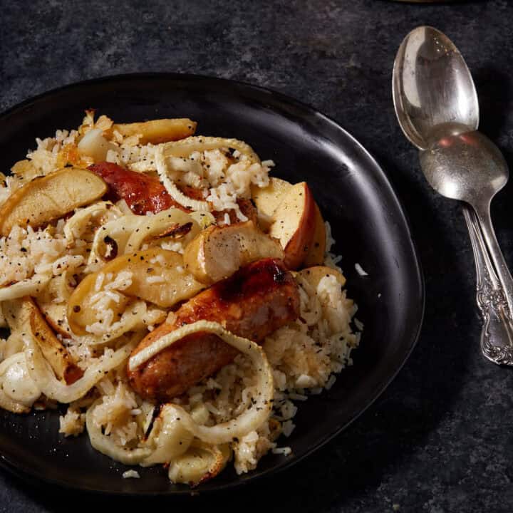 A black platter filled with sausages and apples next to two serving spoons.