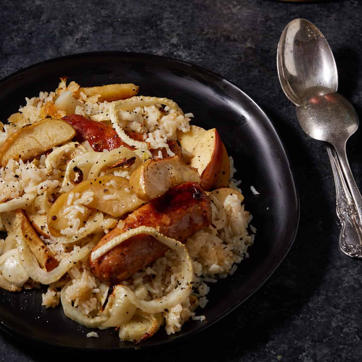A black platter filled with sausages and apples next to two serving spoons.