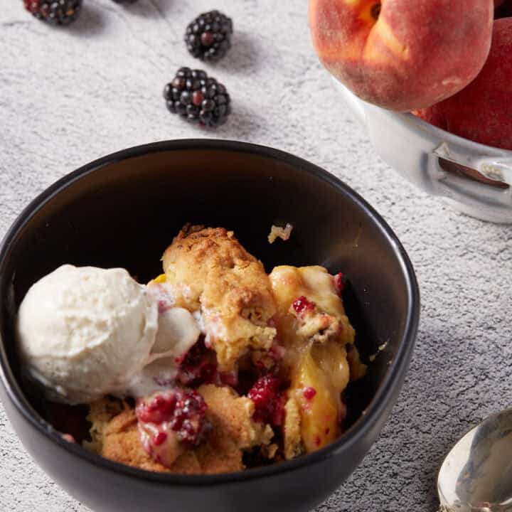 Southern Blackberry Cobbler with peaches in a black bowl with a scoop of ice cream.