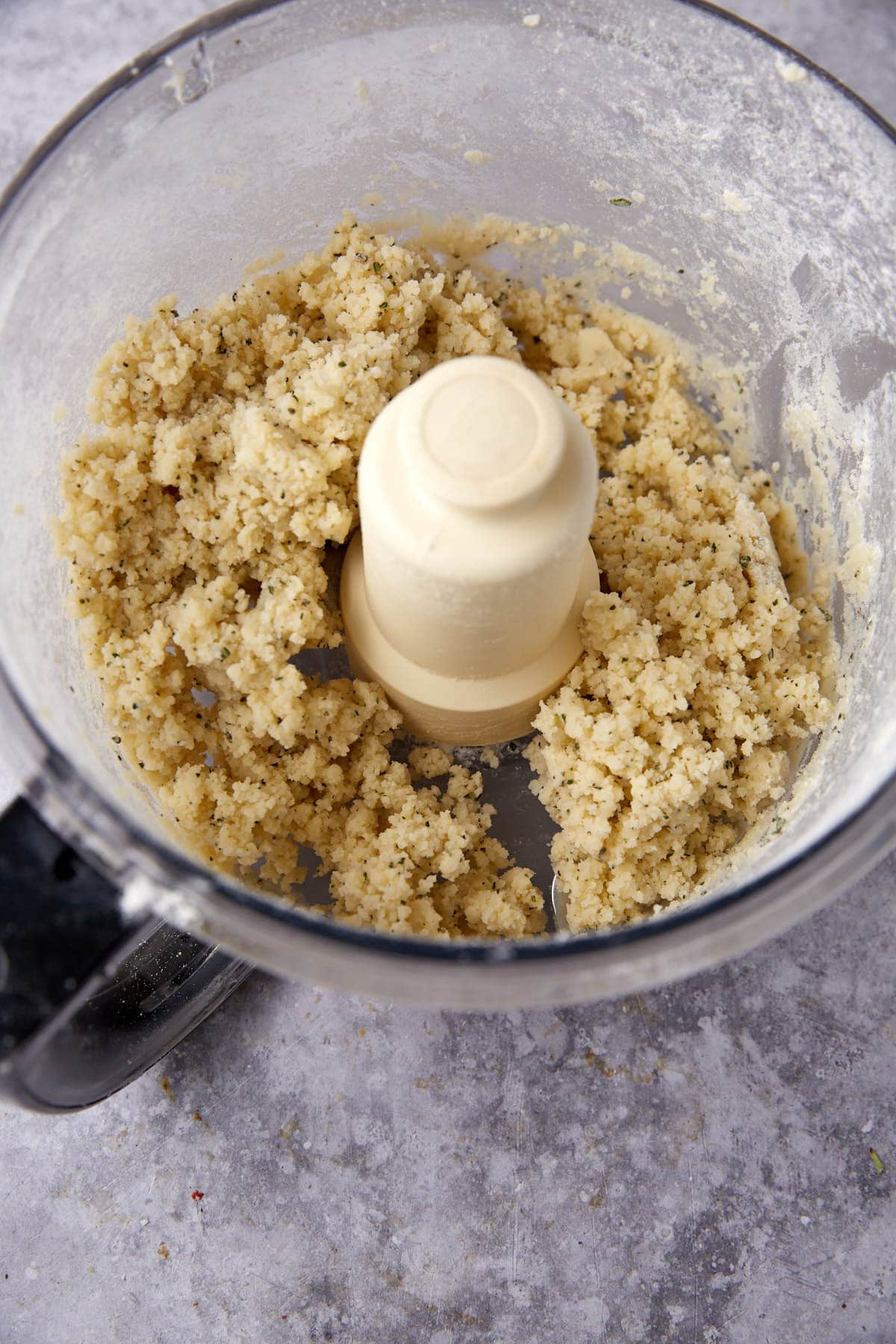 Crumbled dough mixture for cheese crackers inside a food processor bowl on a gray countertop, with small bits sticking to the sides and blade.