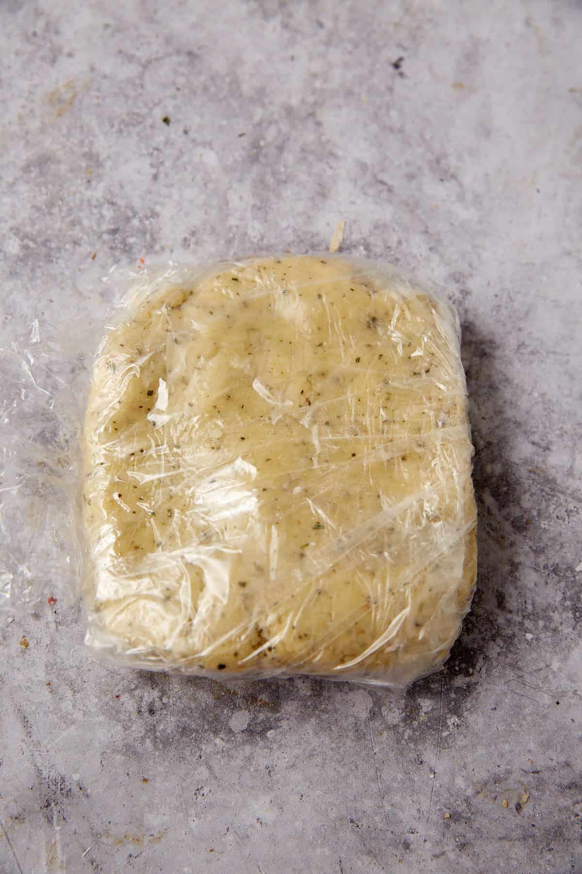 A square-shaped portion of dough with visible herbs, perfect for making cheese crackers, is wrapped in plastic wrap and placed on a lightly textured gray surface.