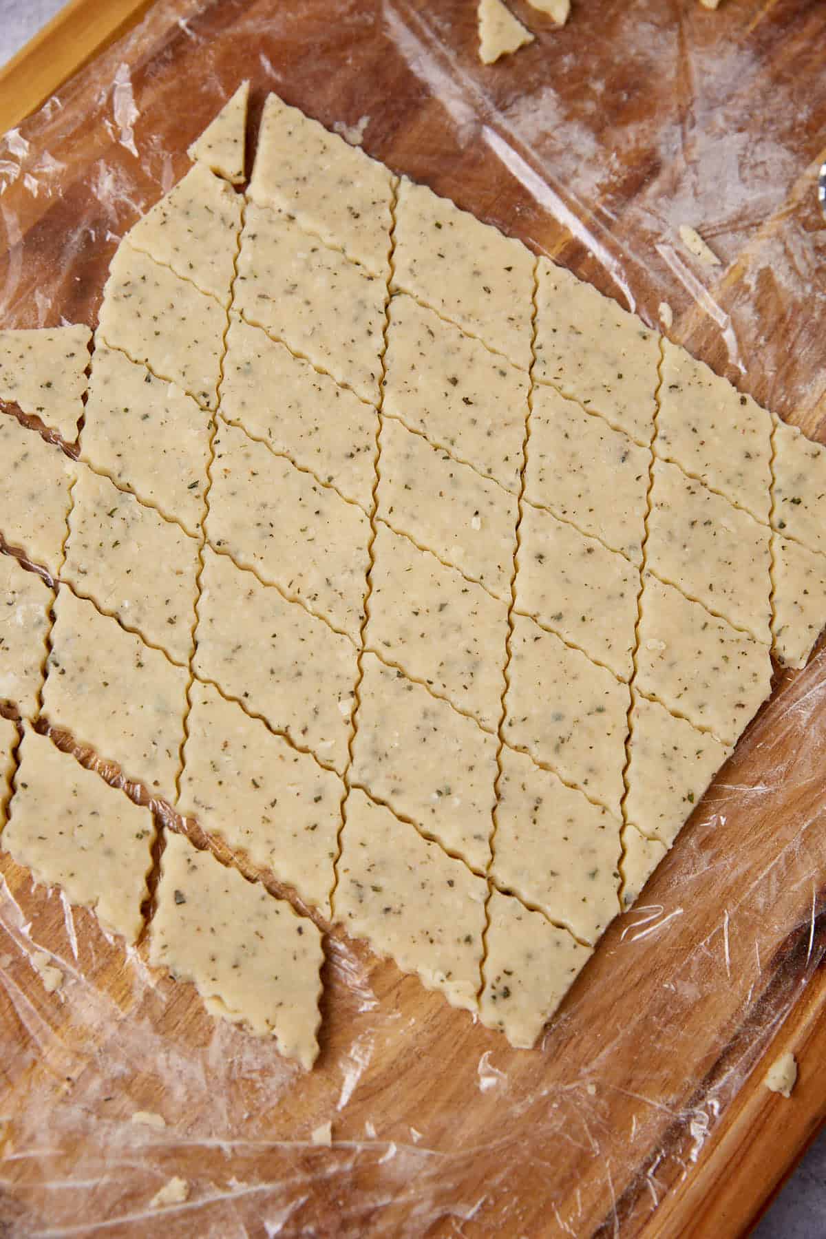 Diamond-shaped cheese cracker dough with herbs, cut into pieces on a wooden board, ready for baking. The speckled dough rests on clear plastic wrap, with scraps and flour scattered around the edges.
