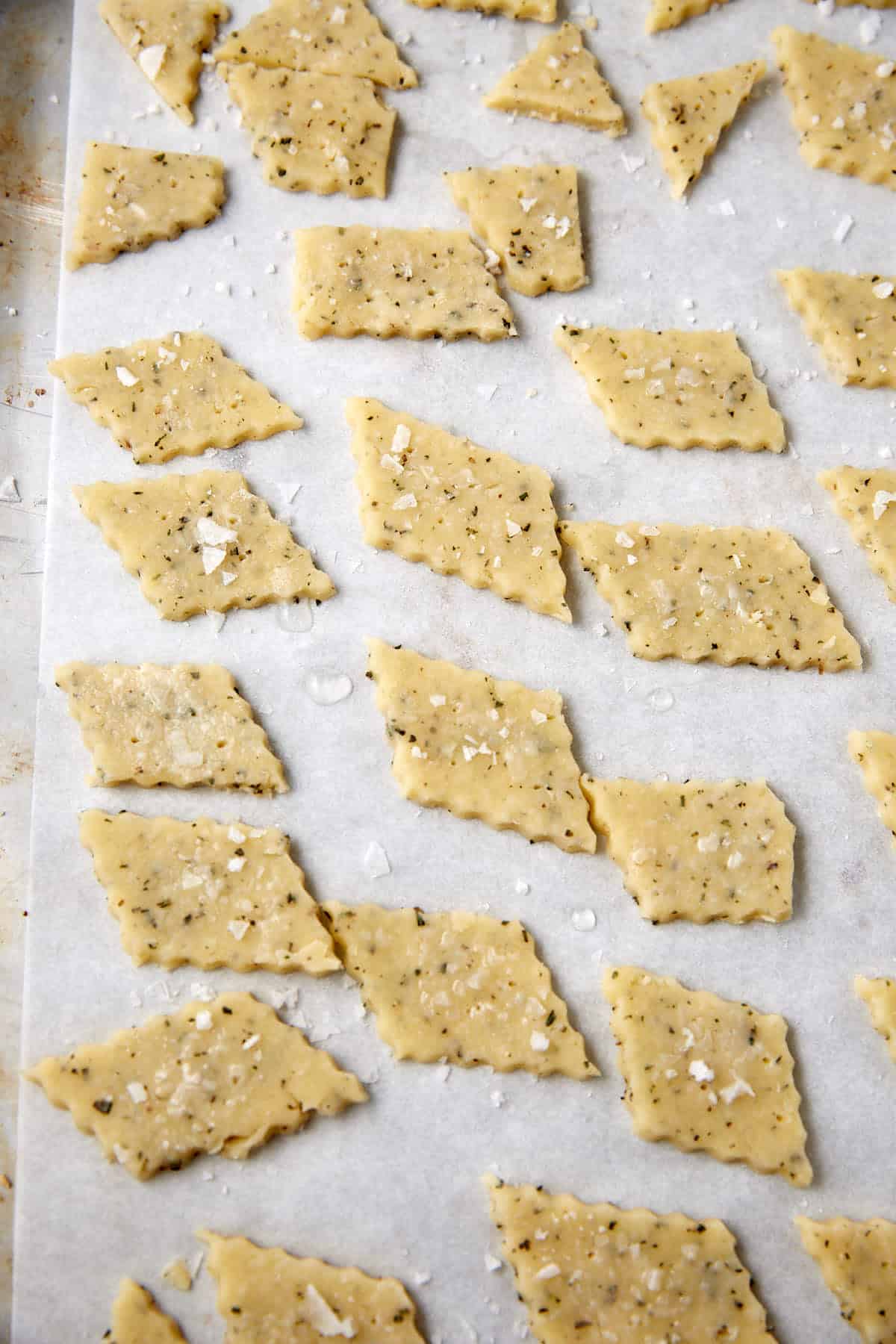 Diamond-shaped pieces of uncooked, herbed cheese cracker dough are arranged on parchment paper, sprinkled with flaky salt, ready to be baked.