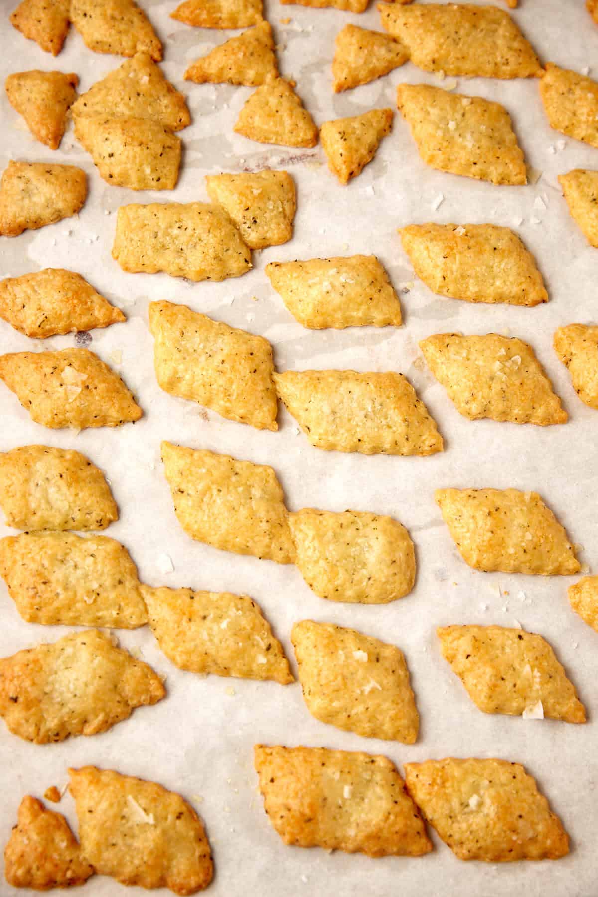 Diamond-shaped cheese crackers are arranged on a sheet of parchment paper. The golden brown cheese crackers, sprinkled with salt, are spaced evenly apart in neat rows.