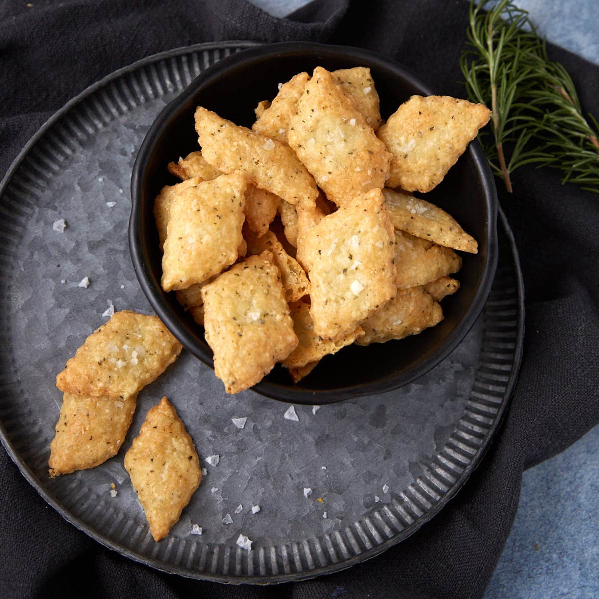 A black bowl filled with diamond-shaped, golden brown cheese crackers sprinkled with salt, placed on a dark metal plate with a few crackers and salt flakes scattered around. A sprig of rosemary lies nearby.