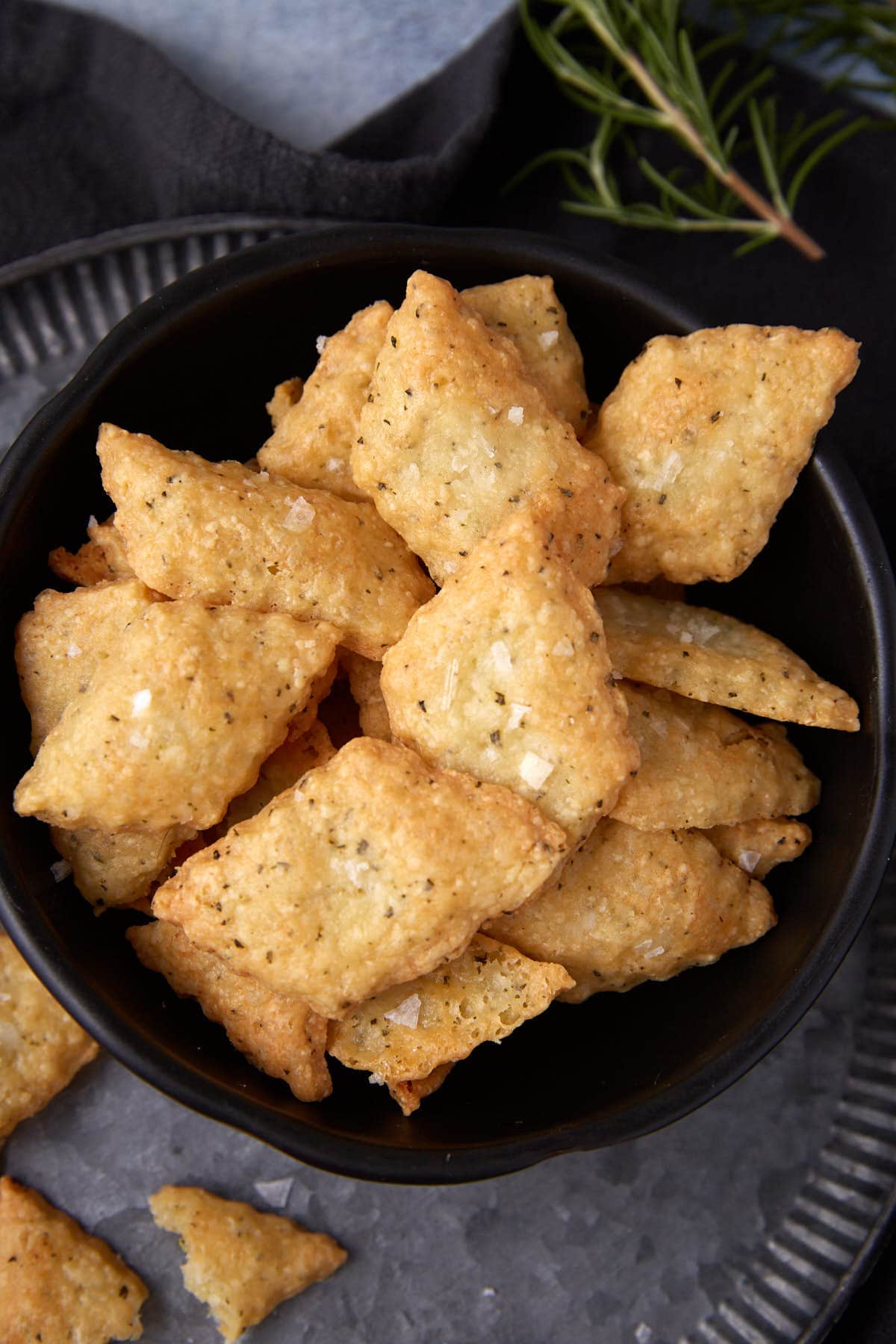 A black bowl filled with golden, crispy, diamond-shaped cheese crackers sprinkled with salt, placed on a metal tray with a sprig of rosemary in the background.