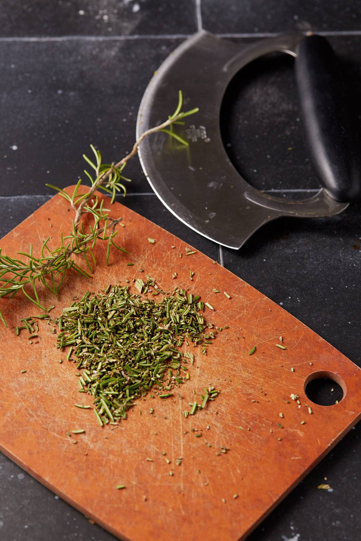 A sprig of fresh rosemary and a pile of chopped rosemary sit on a brown cutting board, next to a mezzaluna knife—perfect for adding bold flavor to homemade cheese crackers on a dark countertop.