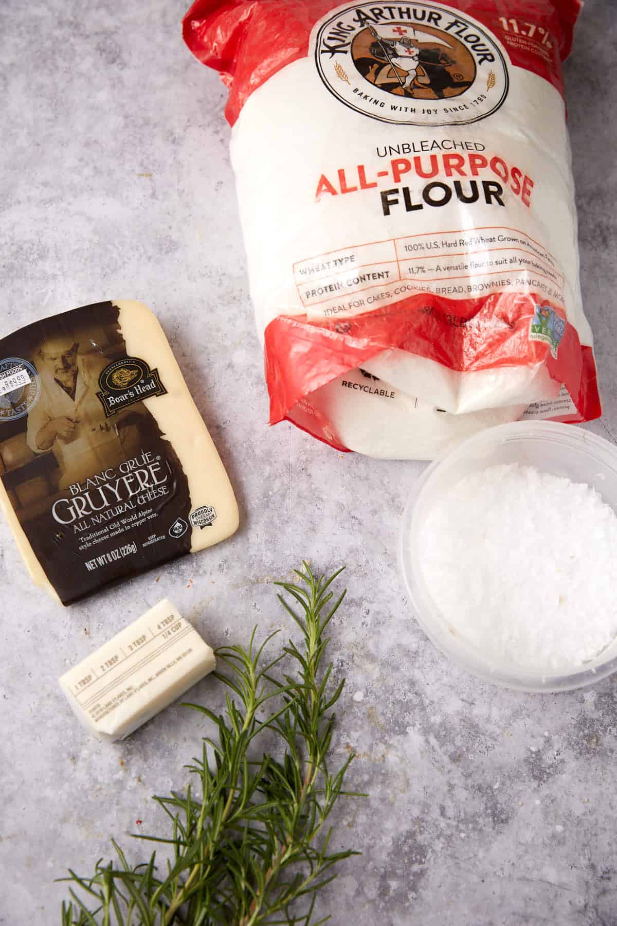 A bundle of fresh rosemary, a stick of butter, a wedge of gruyere cheese, salt, and King Arthur Flour are arranged on a gray countertop—everything you need for delicious homemade cheese crackers.