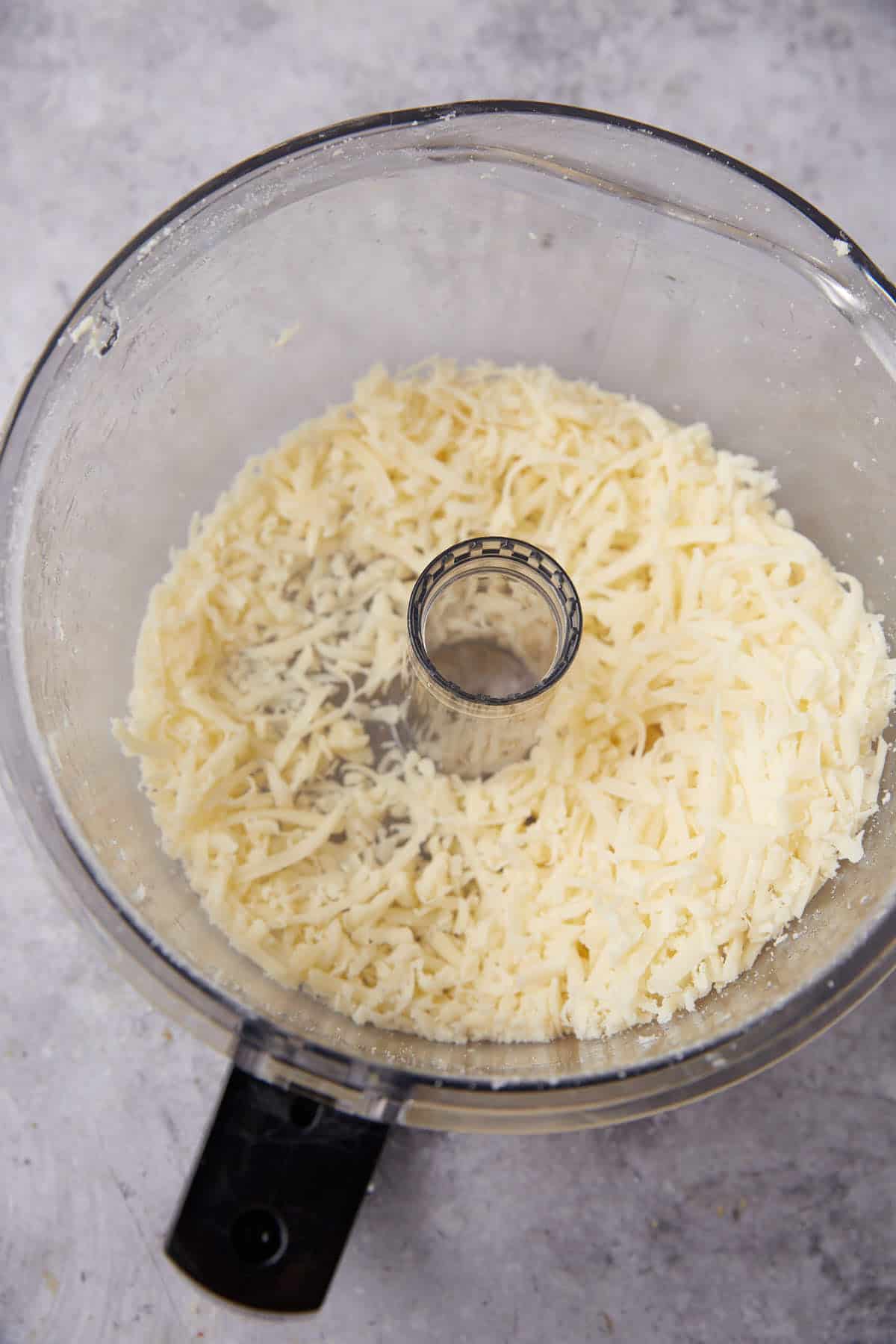 Shredded cheese in a food processor bowl, perfect for making homemade cheese crackers, viewed from above on a light gray surface.