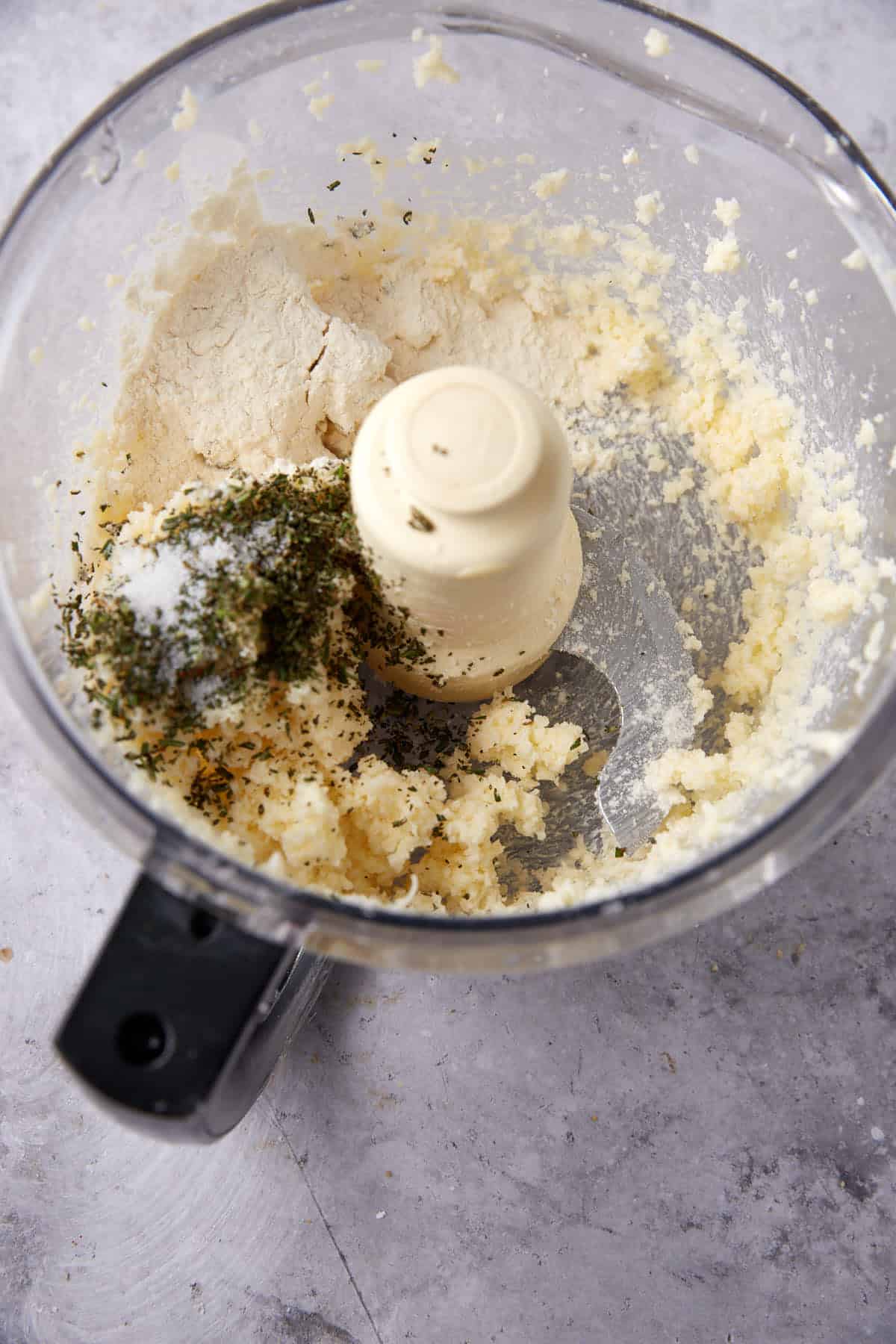 A food processor bowl containing a mixture of flour, creamed butter, cheese crackers, herbs, and salt sits on a gray countertop.