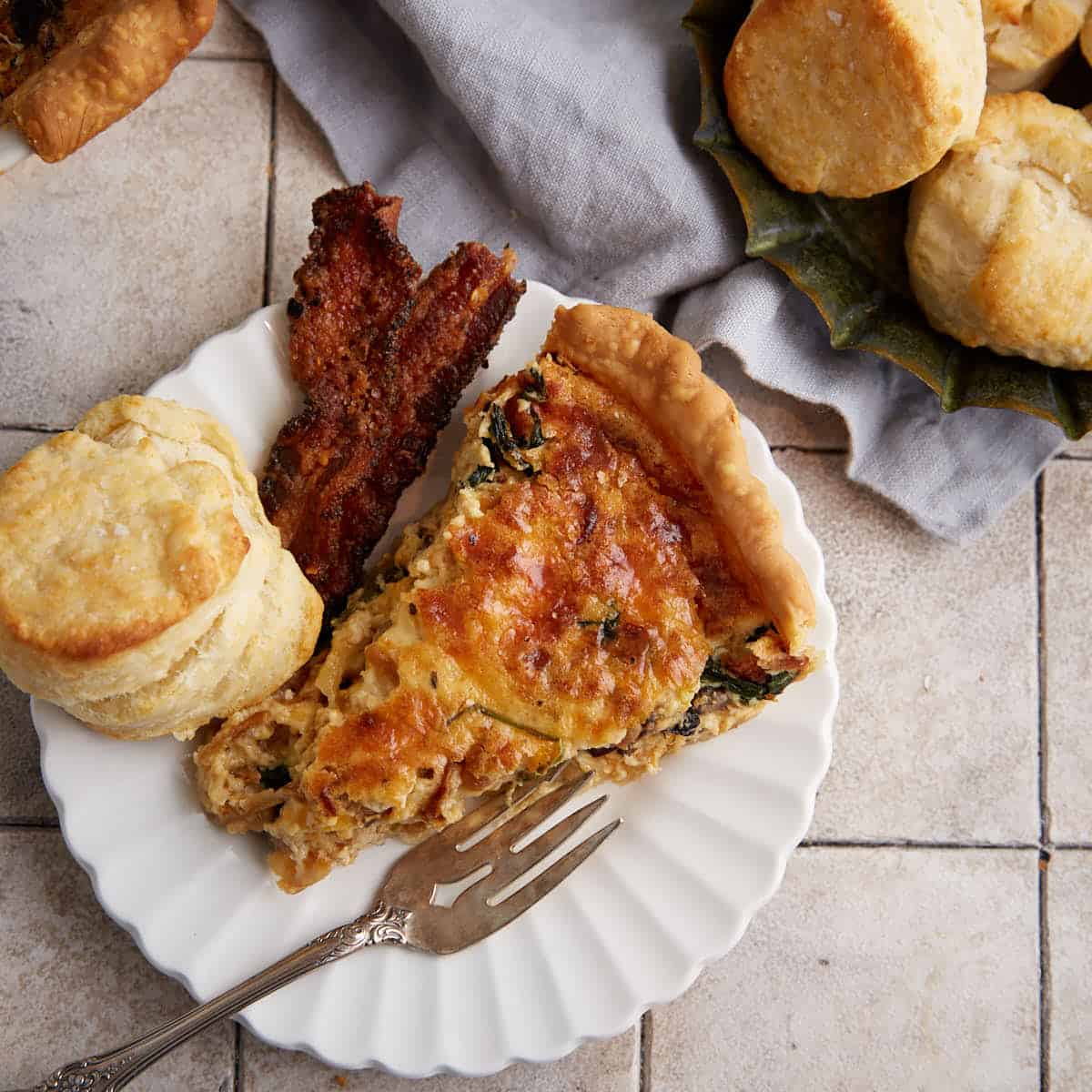 A slice of quiche on a plate with bacon and a biscuit.