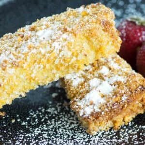 Close-up of two golden French Toast Sticks dusted with powdered sugar on a black plate. In the background, two fresh strawberries are partially visible, adding a touch of color to the presentation.