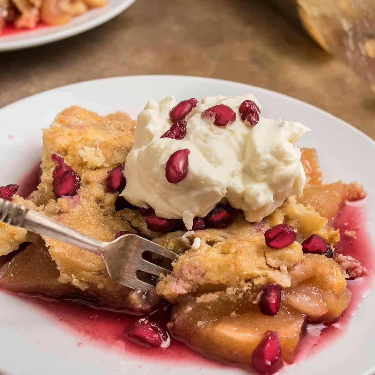 A slice of Apple Pomegranate Cobbler topped with whipped cream and pomegranate seeds sits on a white plate. A fork rests on the dessert, and some fruit filling has spilled onto the plate.