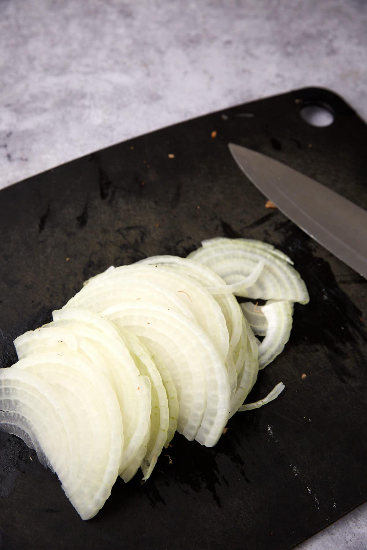 Sliced white onions arranged on a black cutting board next to a kitchen knife, perfect for preparing Green Beans with Bacon and Onions, with a light gray surface in the background.