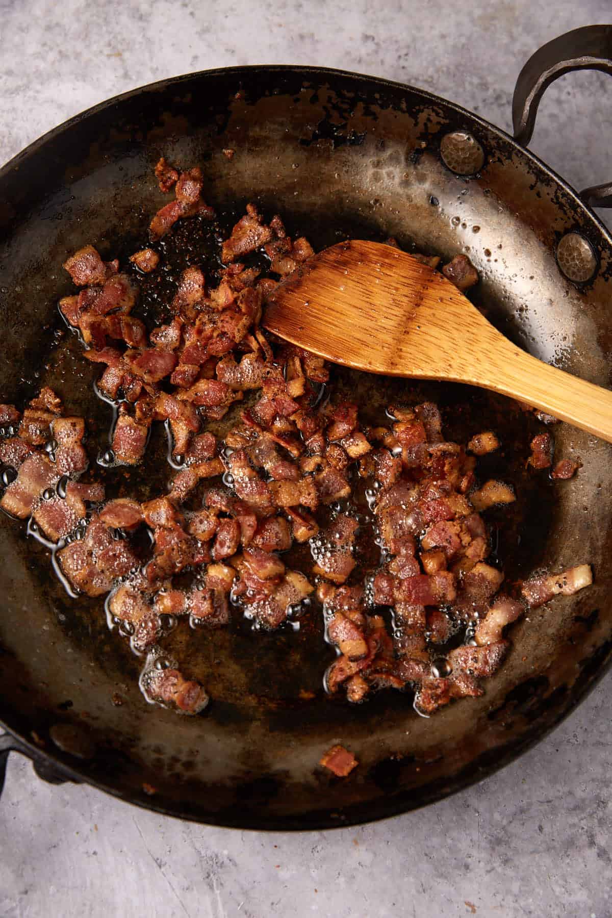 Diced bacon pieces sizzling in a dark, oily skillet with a wooden spatula resting on the pan—perfect for starting Green Beans with Bacon and Onions. The background is a light, textured surface.