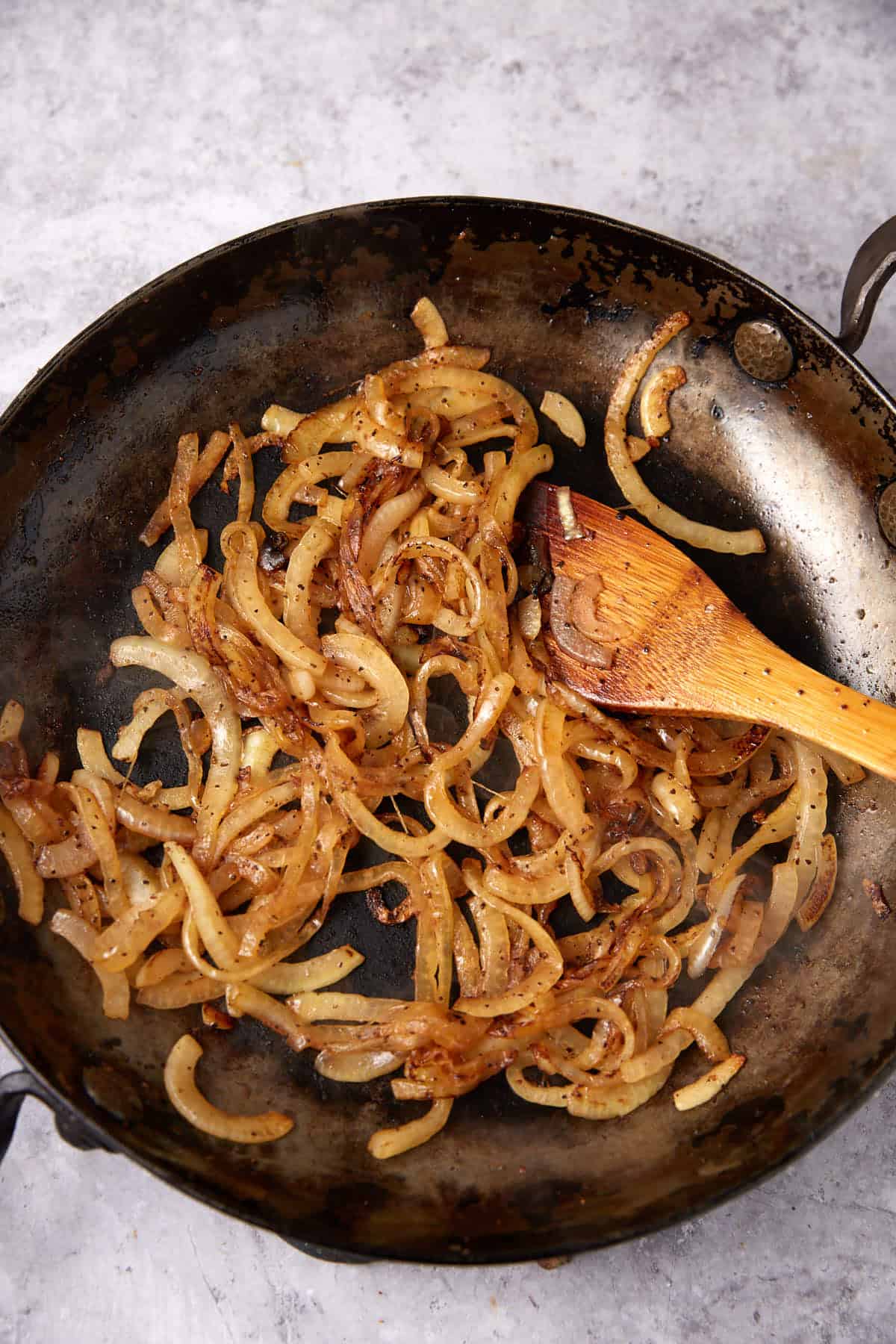 Sliced onions sautéing in a dark skillet with a wooden spoon, lightly browned and seasoned—perfect for pairing with Green Beans with Bacon and Onions on a gray surface.