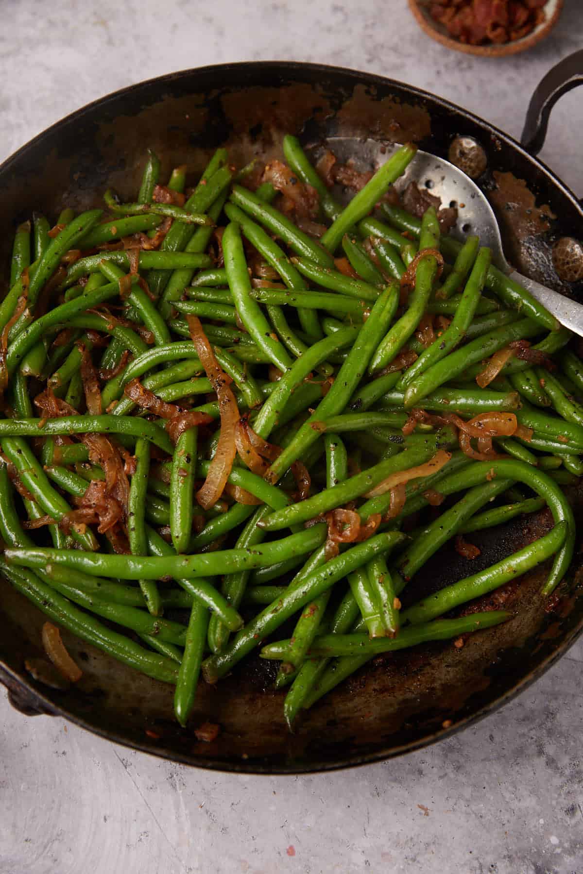 A skillet filled with Green Beans with Bacon and Onions, featuring sautéed green beans and caramelized onions, garnished with savory bacon bits, with a metal serving spoon resting on the side.