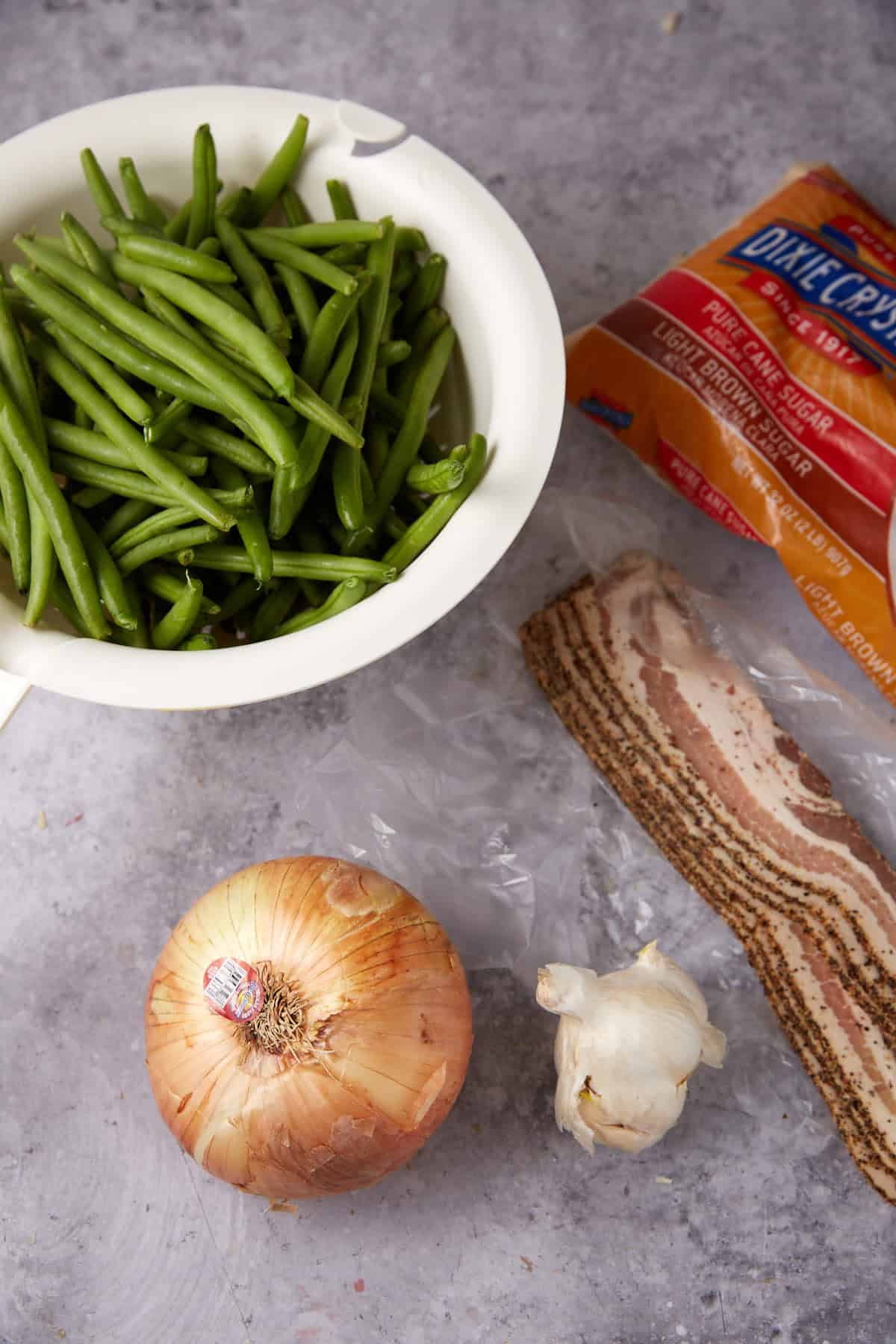 A bowl of fresh green beans, a bag of brown sugar, thick slices of bacon, a yellow onion, and a bulb of garlic arranged on a gray countertop—everything you need for Green Beans with Bacon and Onions.