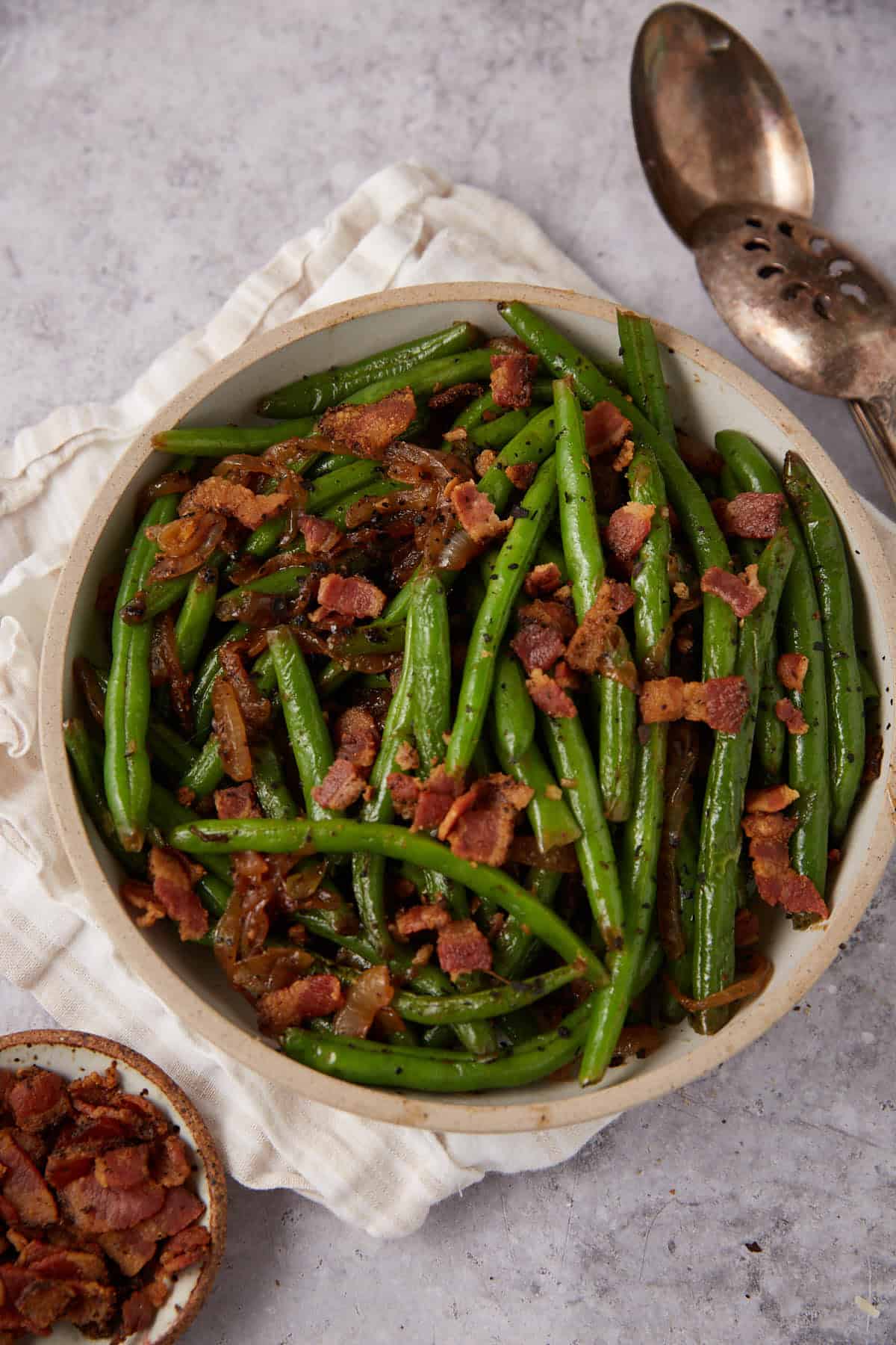 A bowl of Green Beans with Bacon and Onions, topped with crispy bacon pieces and caramelized onions, sits on a light cloth napkin with serving utensils. A small dish of extra bacon crumbles is nearby for added flavor.