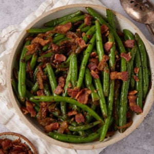 A bowl of Green Beans with Bacon and Onions features sautéed green beans topped with crispy bacon pieces and caramelized onions, set on a light-colored cloth with a serving spoon nearby.