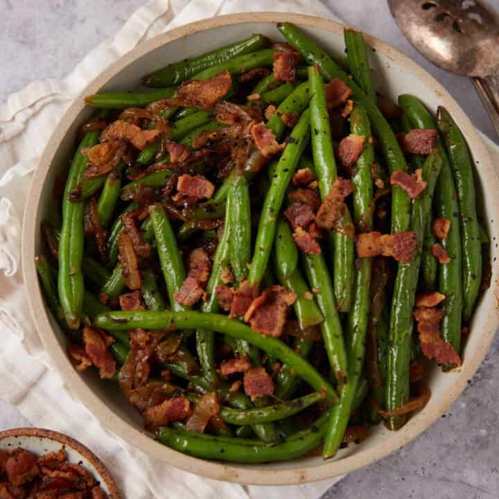 A bowl of Green Beans with Bacon and Onions features sautéed green beans topped with crispy bacon pieces and caramelized onions, set on a light-colored cloth with a serving spoon nearby.