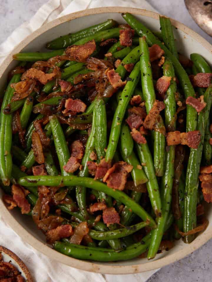A bowl of Green Beans with Bacon and Onions features sautéed green beans topped with crispy bacon pieces and caramelized onions, set on a light-colored cloth with a serving spoon nearby.