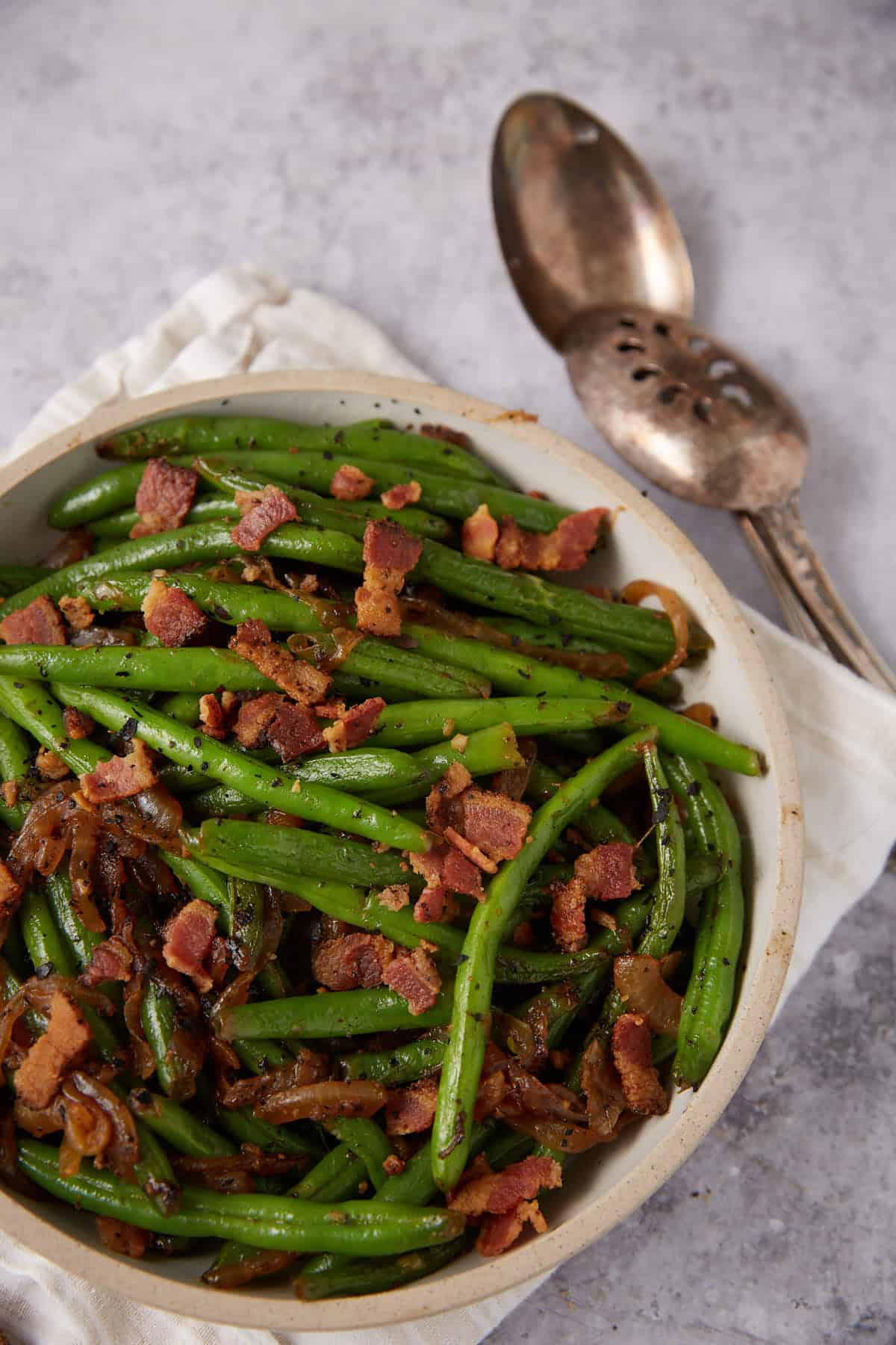 A bowl of Green Beans with Bacon and Onions sits on a white cloth, with crumbled bacon on top and a serving spoon and slotted spoon beside it on a gray surface.