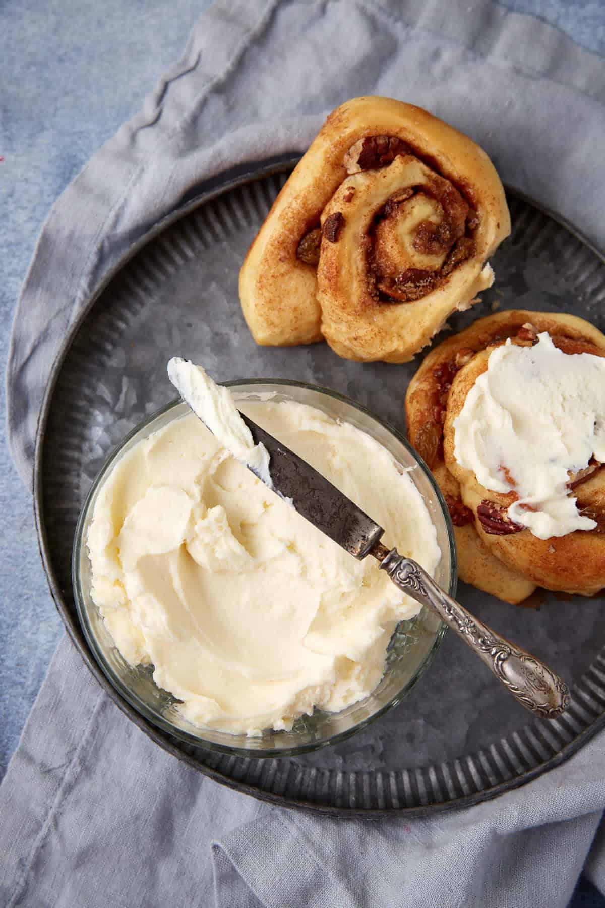 Two cinnamon rolls on a plate with a bowl of cream cheese frosting.