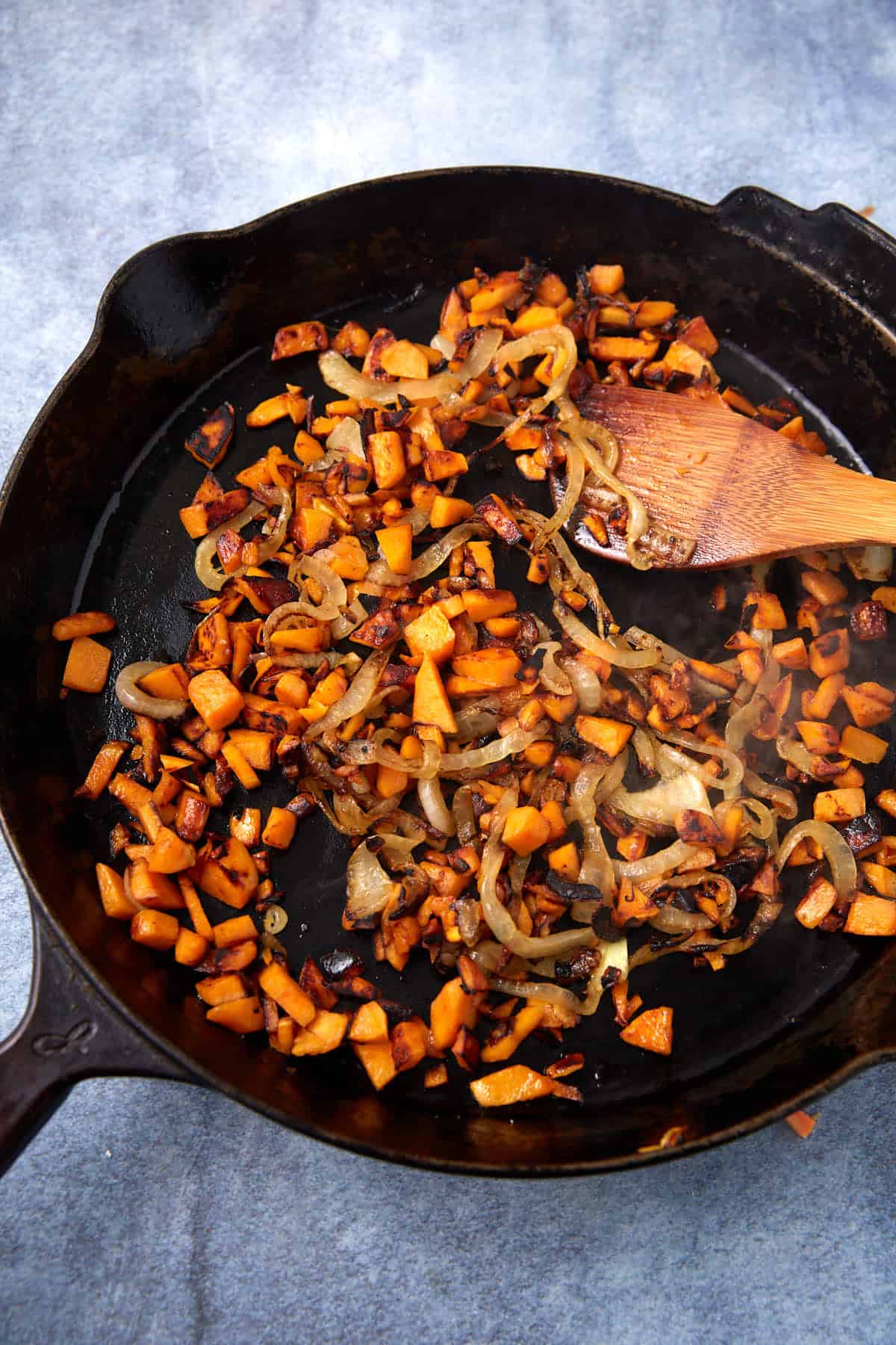 A cast iron skillet with sautéed diced sweet potatoes and sliced onions, partially browned, being stirred with a wooden spatula on a blue countertop—perfect as a hearty filling for Breakfast Burritos.