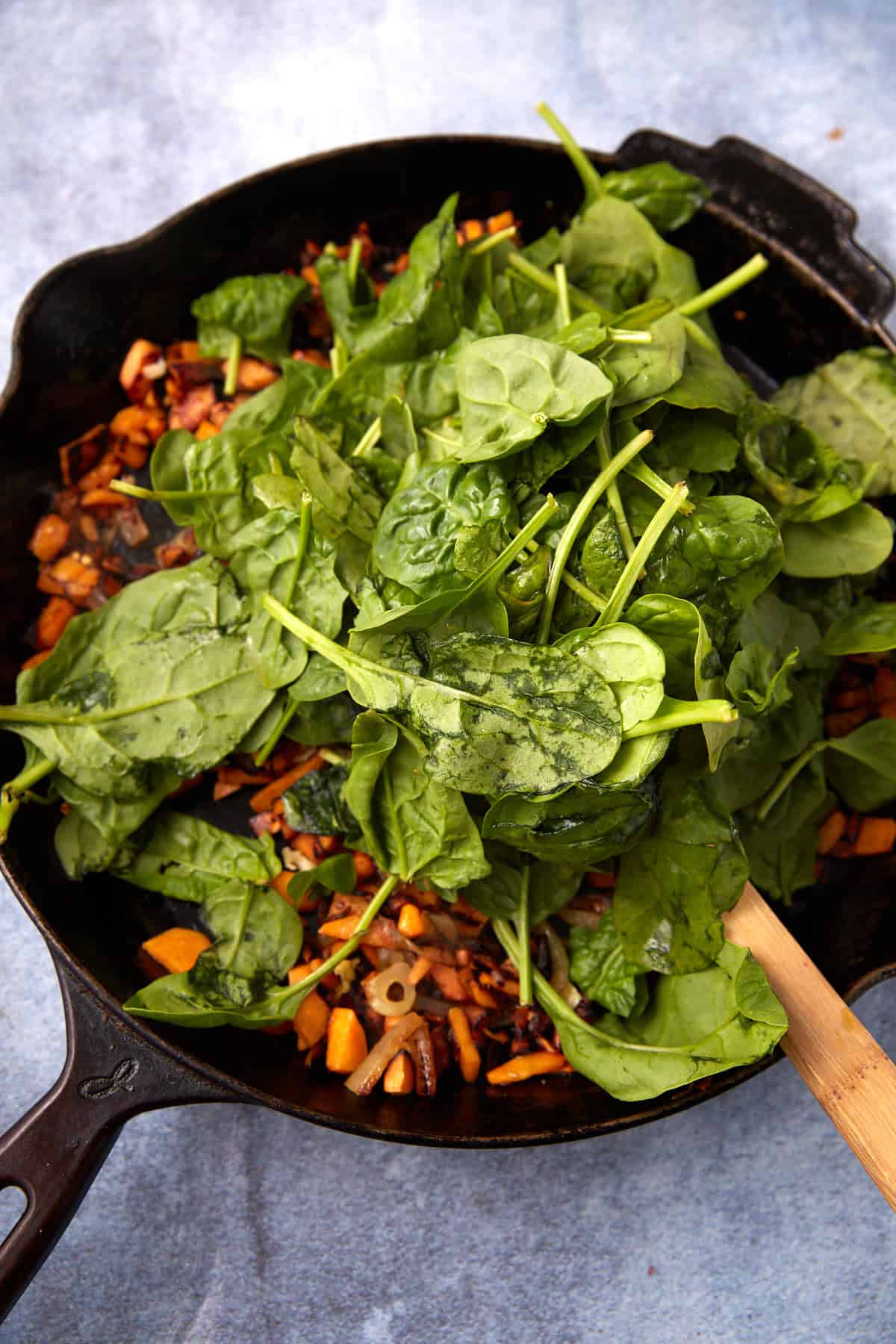 A cast iron skillet filled with fresh spinach leaves atop sautéed chopped onions and carrots, perfect for adding to breakfast burritos, with a wooden spatula resting on the side. The skillet is placed on a light blue surface.