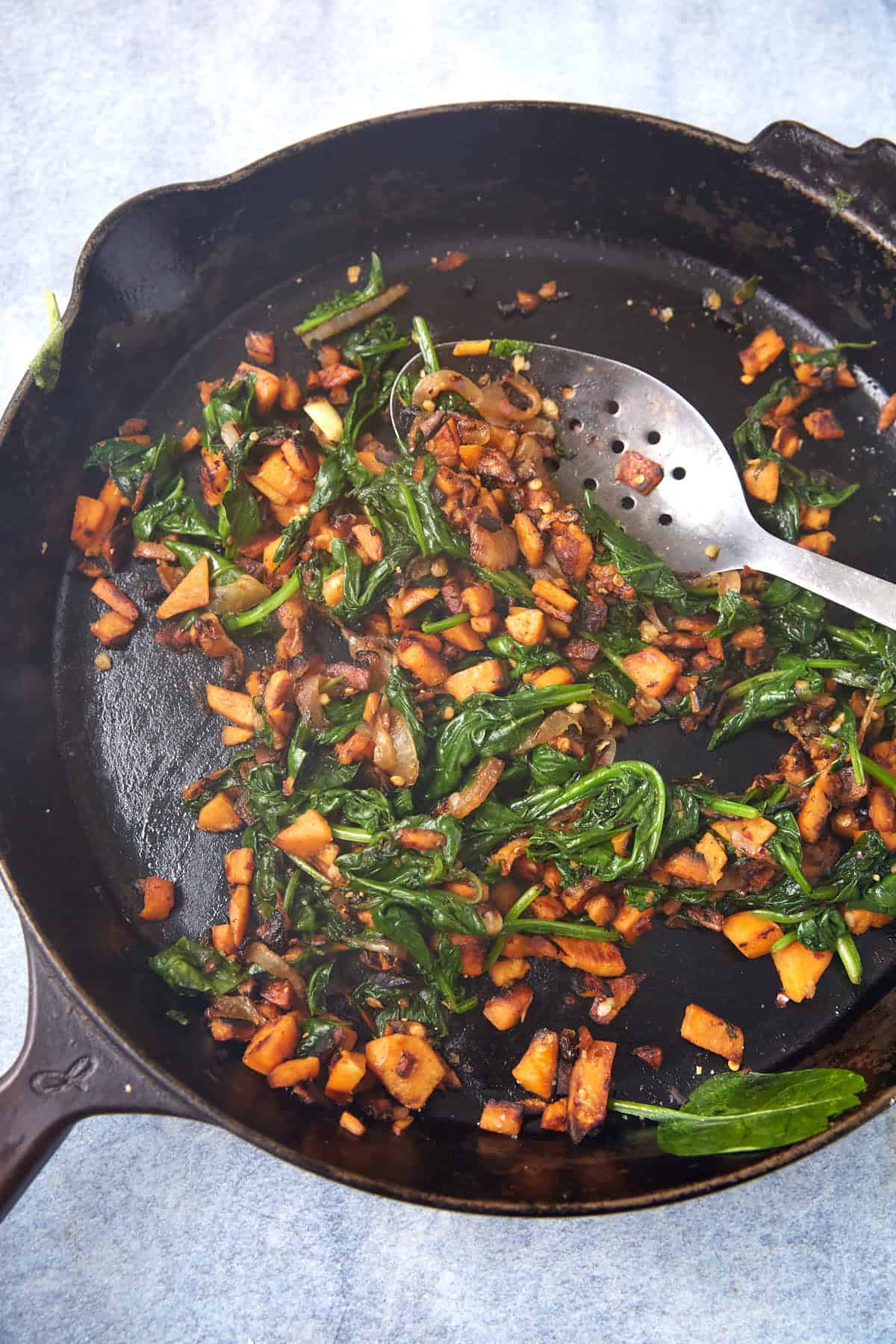 A cast-iron skillet contains sautéed greens, diced sweet potatoes, and onions—perfect fillings for Breakfast Burritos—with a metal slotted spoon resting inside the pan on a light blue surface.