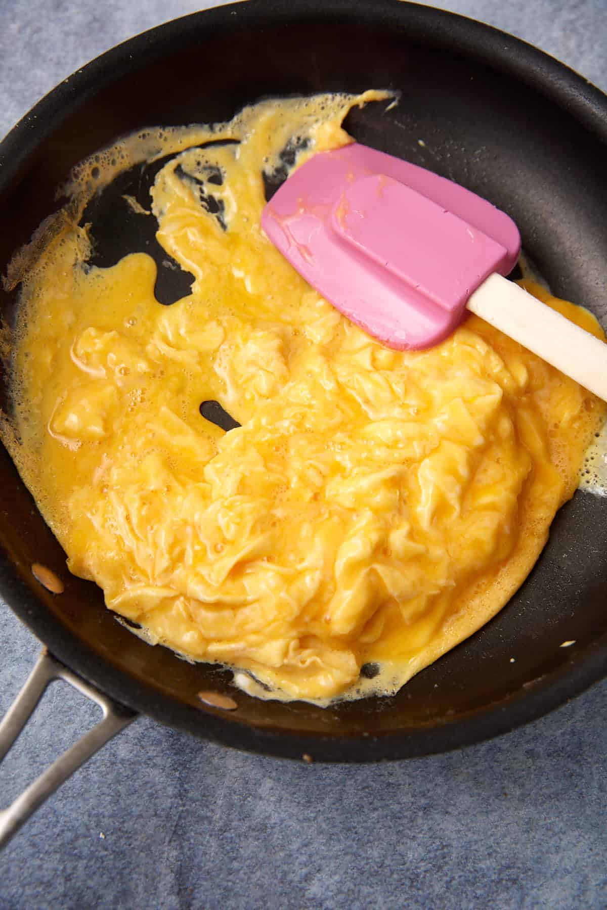 A nonstick frying pan with scrambled eggs being cooked, partially set, and a purple spatula stirring them on a blue surface—perfect for making tasty Breakfast Burritos.