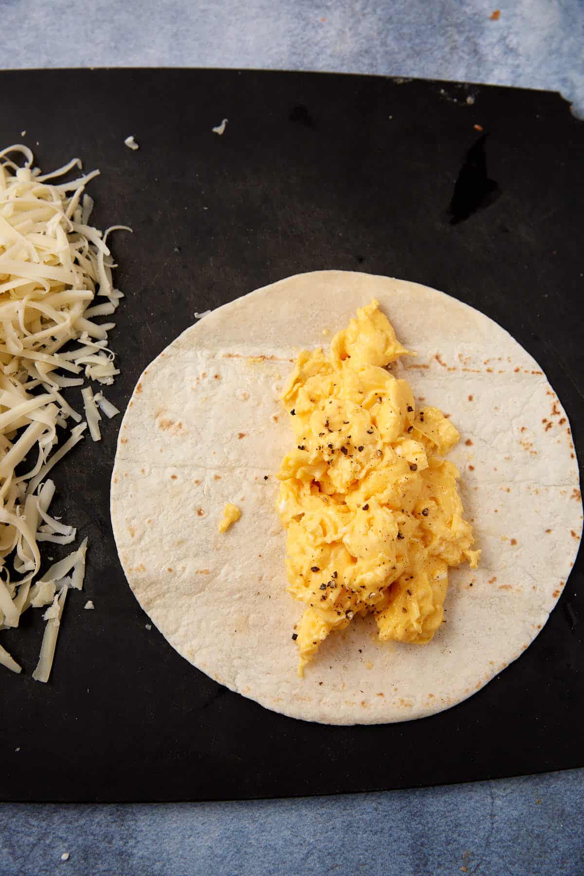 A flour tortilla on a black surface is topped with scrambled eggs and cracked black pepper; a pile of shredded cheese sits beside it, ready to be added—perfect for crafting delicious Breakfast Burritos.