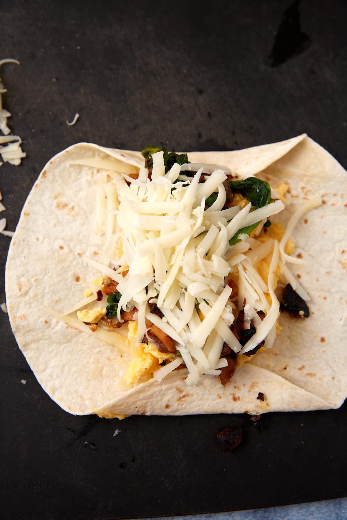 A Breakfast Burrito with scrambled eggs, sautéed vegetables, and shredded cheese sits on a dark surface, ready to be folded, with extra cheese scattered nearby.