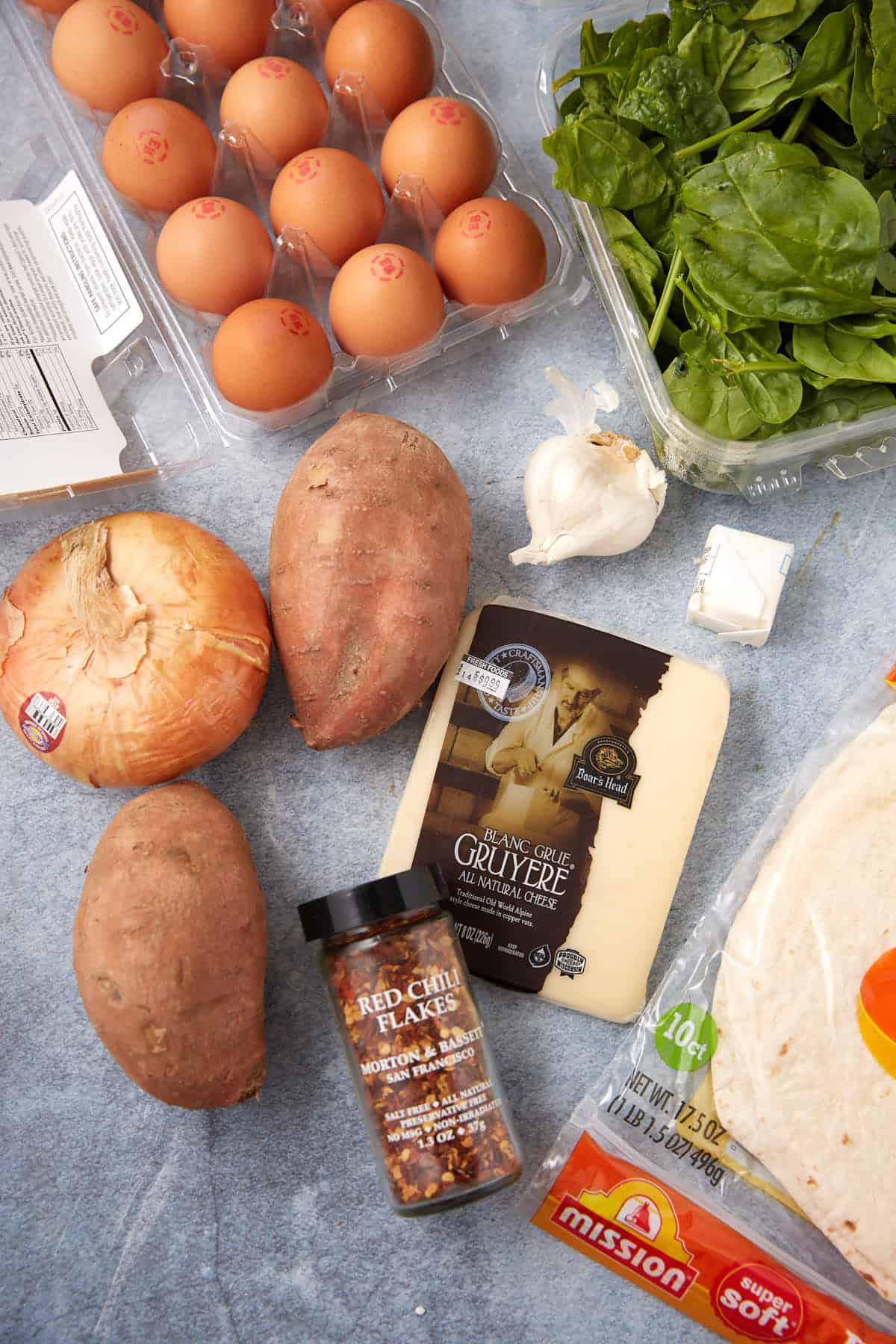 A flat lay of Breakfast Burritos ingredients on a gray surface: eggs, spinach, sweet potatoes, onions, Gruyère cheese, garlic, butter, red chili flakes, and flour tortillas.