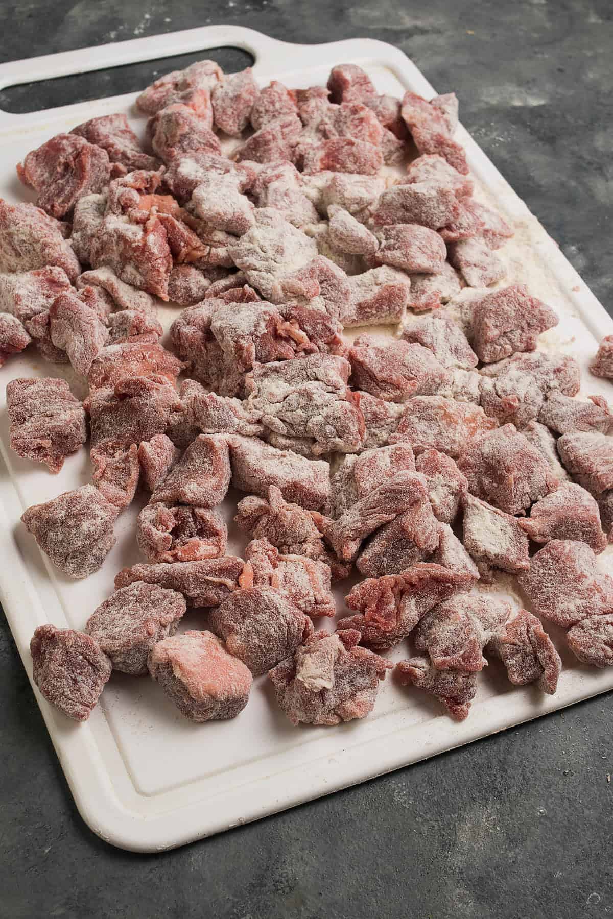 Chunks of raw beef, ready for a classic beef and Guinness pie, are coated in flour and spread out on a white cutting board placed on a gray countertop.