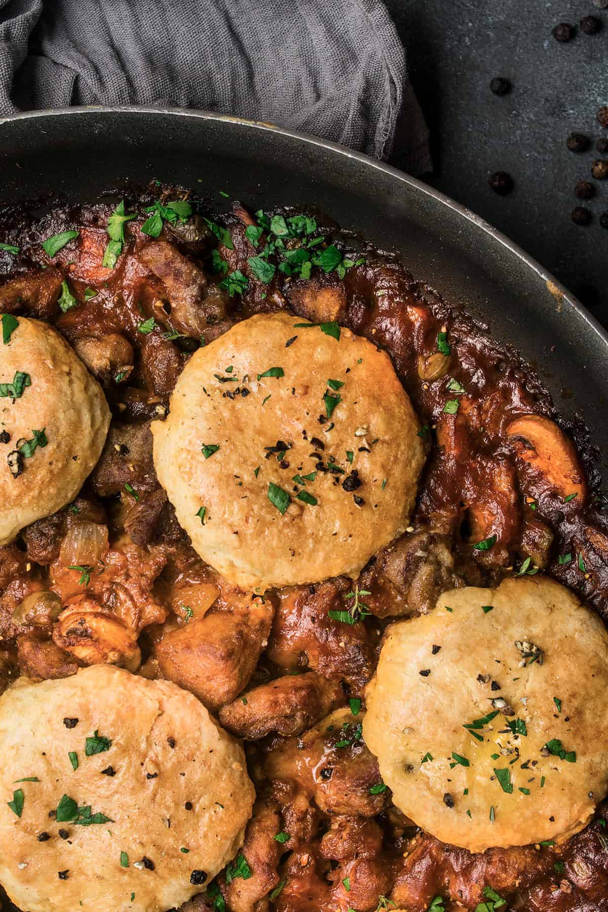 A skillet filled with a rich beef and Guinness pie stew of meat and vegetables, topped with golden, flaky biscuits and garnished with chopped parsley. A gray cloth napkin is partially visible in the corner.