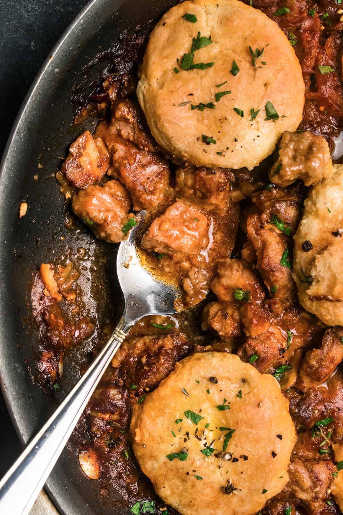 A close-up of a skillet filled with savory beef and Guinness pie stew and golden-brown biscuits, garnished with herbs, with a spoon scooping up some of the hearty filling.