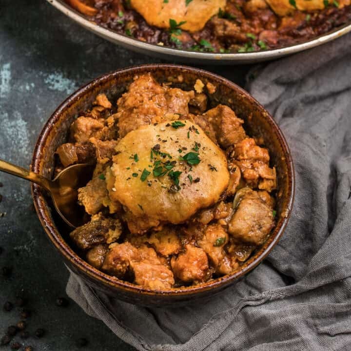 A rustic bowl filled with a hearty beef and Guinness pie-inspired meat and vegetable stew, topped with a golden, flaky biscuit and garnished with chopped herbs. A gold spoon rests inside the bowl, and a gray cloth napkin lies nearby.