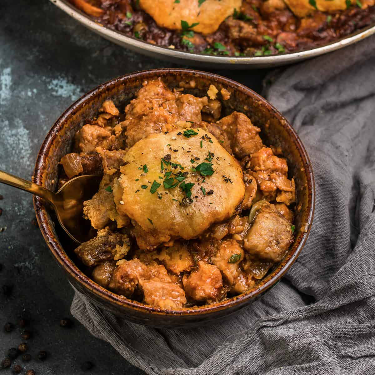 A rustic bowl filled with a hearty beef and Guinness pie-inspired meat and vegetable stew, topped with a golden, flaky biscuit and garnished with chopped herbs. A gold spoon rests inside the bowl, and a gray cloth napkin lies nearby.