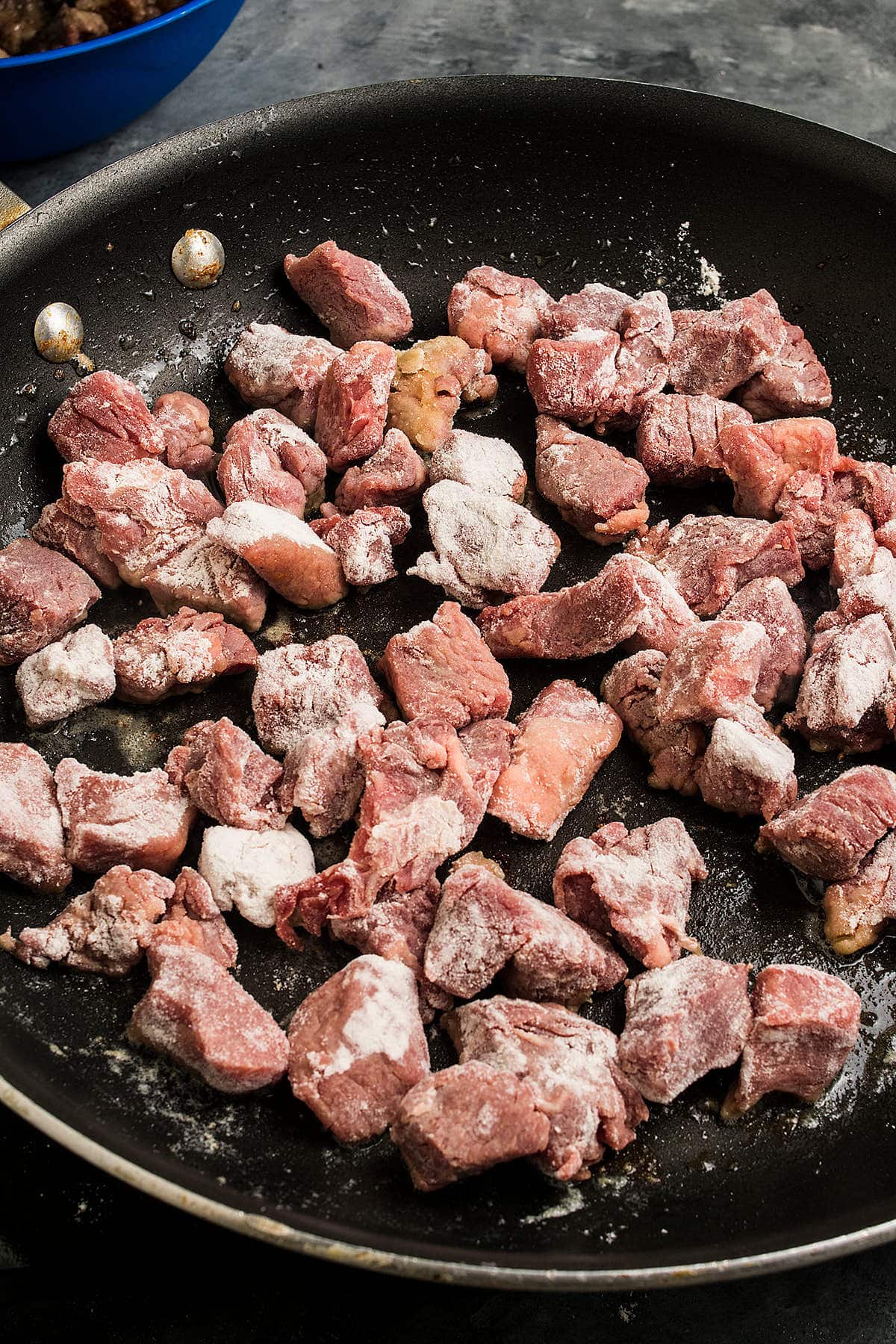 Chunks of raw beef, perfect for a hearty beef and guinness pie, are coated in flour and spread out in a black frying pan, ready to be cooked. The background is slightly blurred, revealing a hint of a blue bowl.