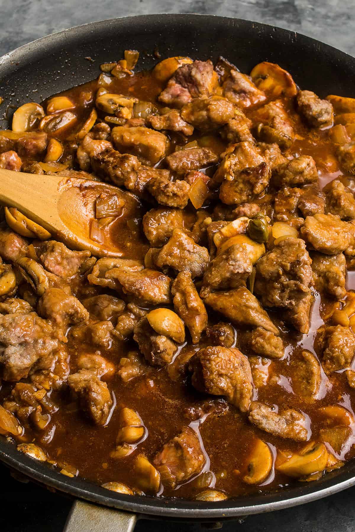A skillet filled with beef strips cooked in a rich brown sauce with sliced mushrooms, reminiscent of the hearty flavors found in classic beef and Guinness pie, being stirred with a wooden spoon.