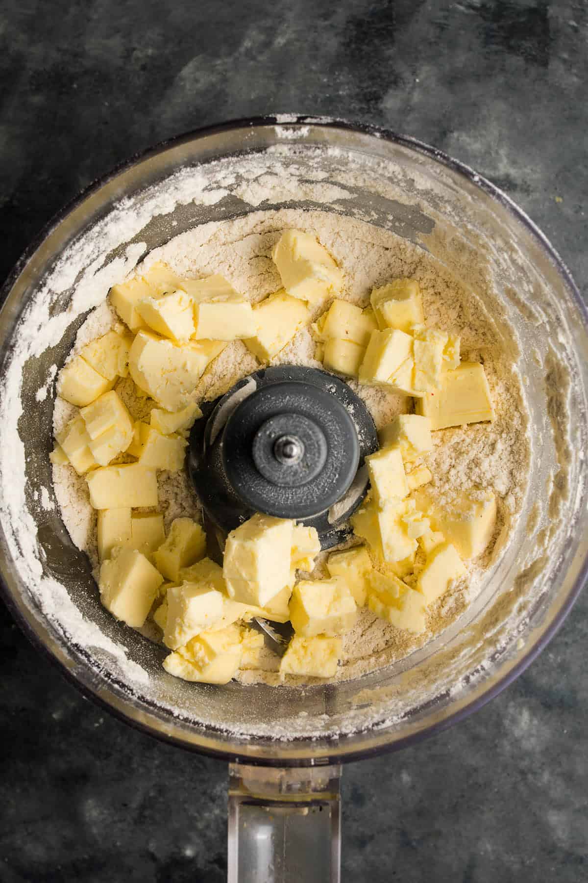 A food processor bowl containing flour and cubes of butter, ready to be mixed for a beef and guinness pie, viewed from above on a dark countertop.