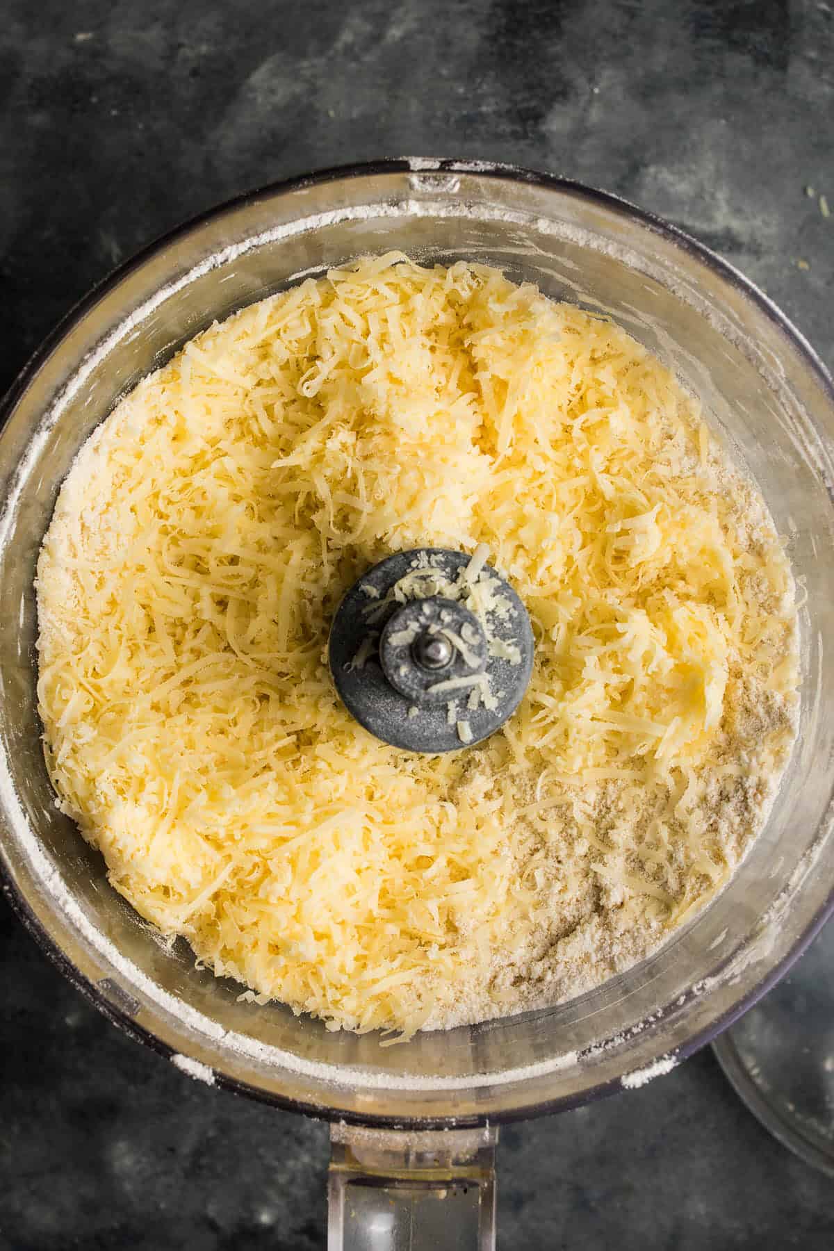 A food processor bowl filled with shredded cheese and flour mixture, viewed from above on a dark countertop, ready to be blended for a savory beef and Guinness pie.