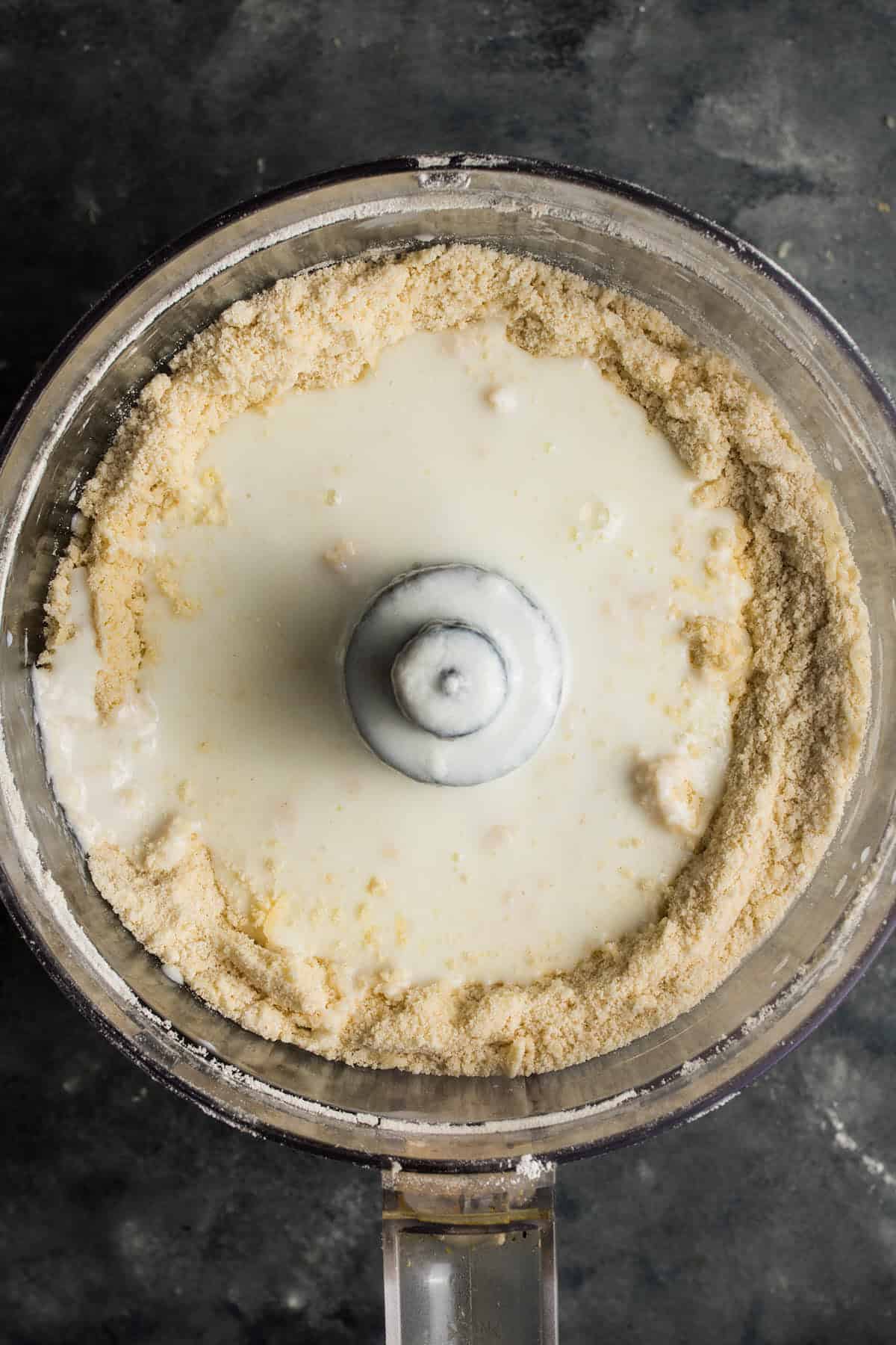 A food processor bowl containing a mixture of flour and a pool of milk, ready to be blended for a delicious beef and Guinness pie, viewed from above on a dark surface.