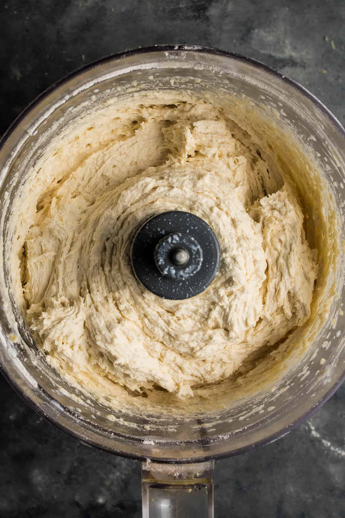 Overhead view of thick bread dough being mixed in a food processor, similar to preparing crust for a hearty beef and Guinness pie, with the blade visible in the center and dough stuck to the sides of the bowl.