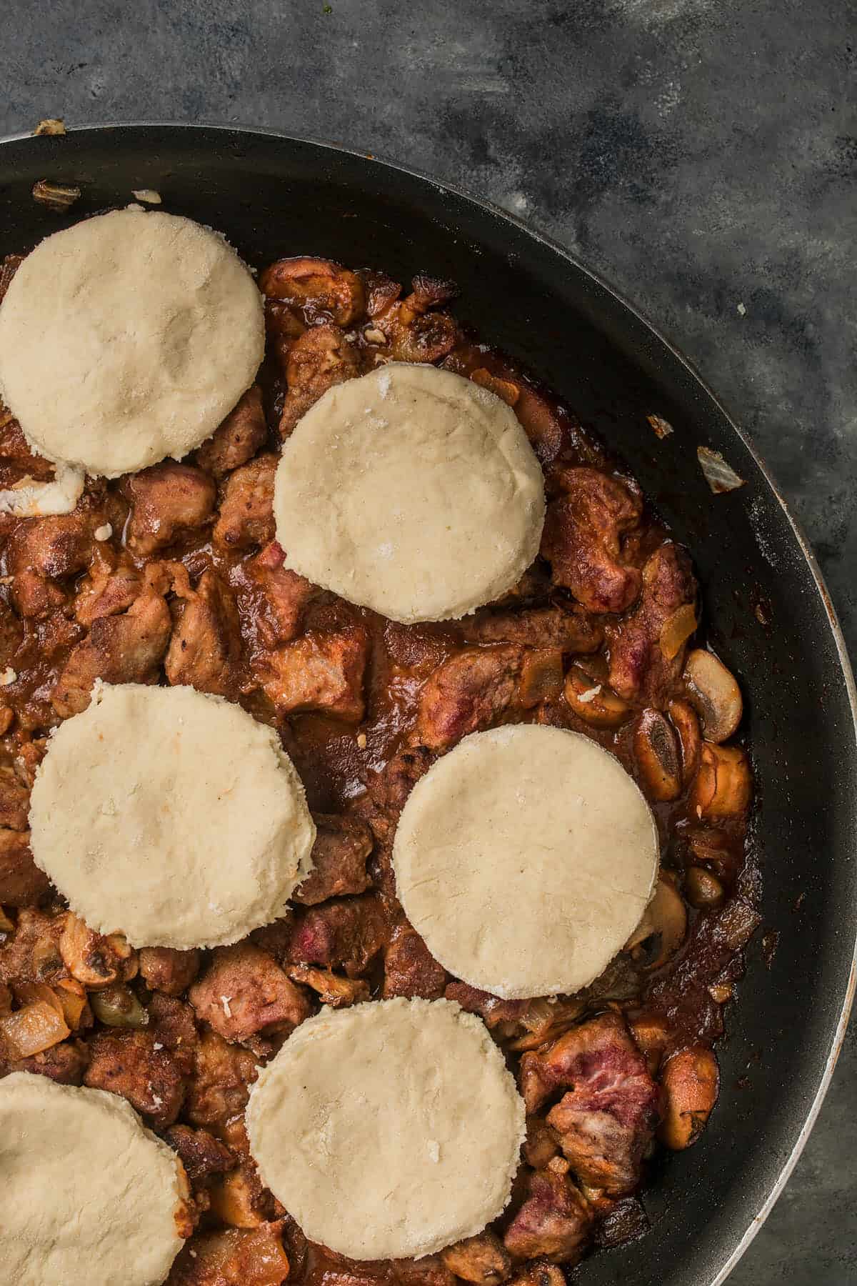 A skillet filled with a rich, chunky beef and Guinness pie stew of meat and vegetables, topped with six round, uncooked biscuit dough pieces, ready to be baked; the background is a dark countertop.
