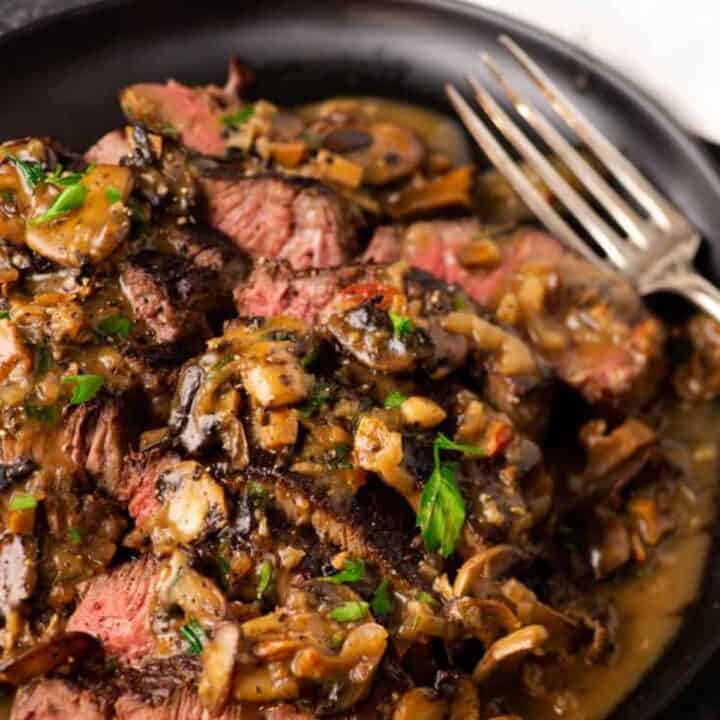 A plate of Steak Marsala topped with a rich mushroom sauce, garnished with chopped parsley. A fork rests on the side of the dish. The steak is sliced, revealing a medium-rare interior. The overall presentation is appetizing and savory.