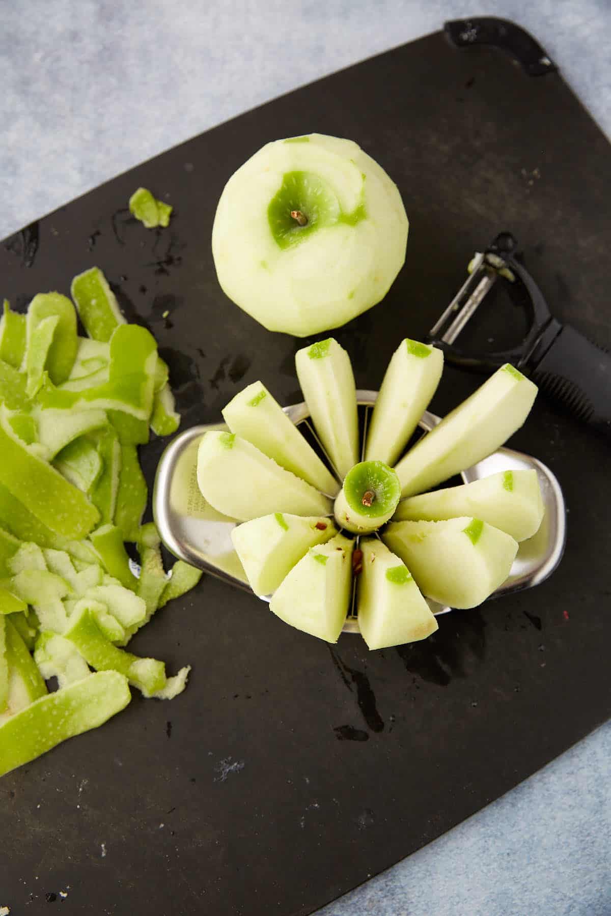 A peeled green apple is being sliced and cored with an apple cutter on a black cutting board, ready for the best Dutch apple pie recipe. Green apple peels and a black peeler lie nearby.